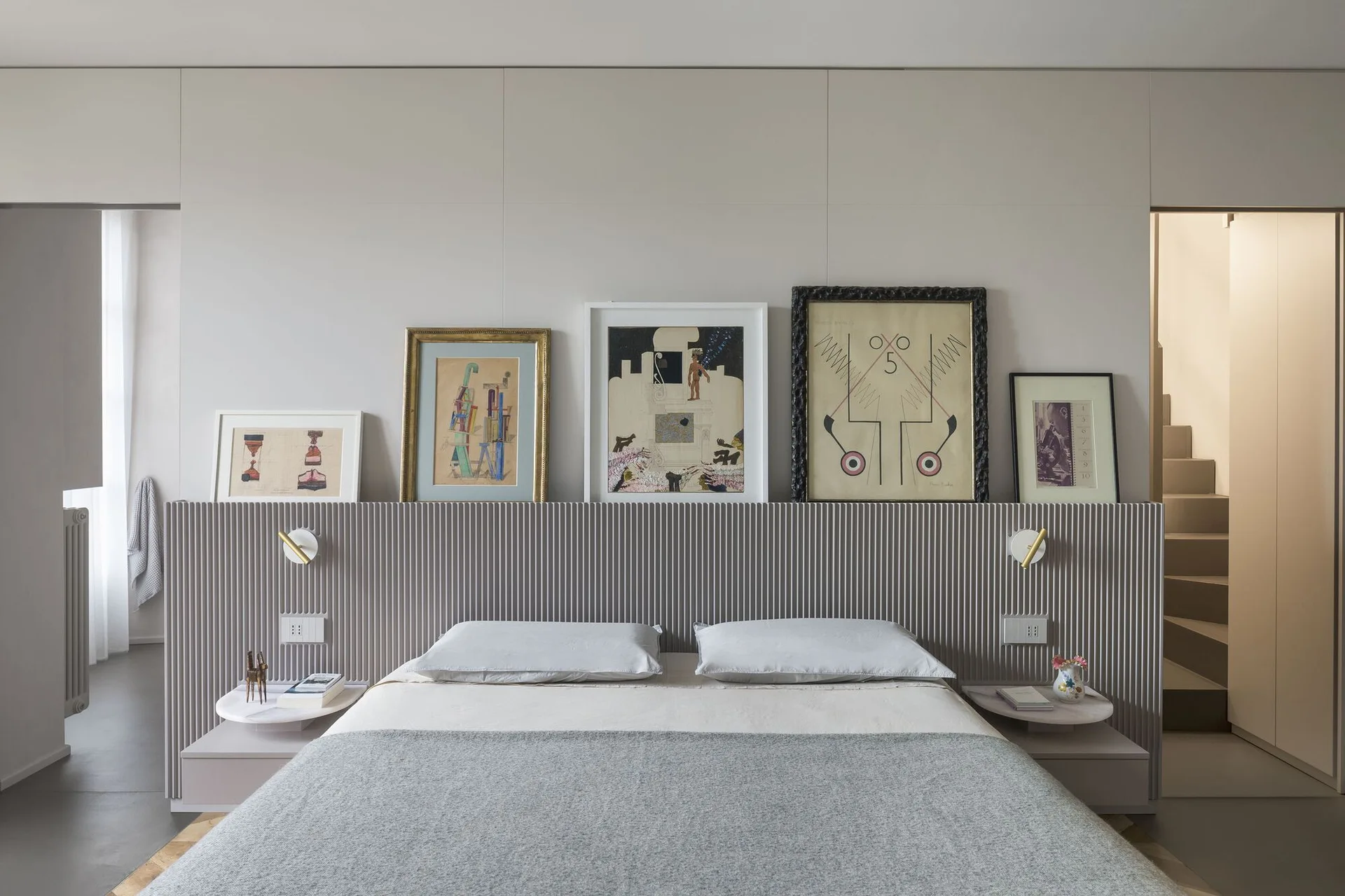 A minimalist grey bedroom features a bed with a wide, fluted headboard that displays framed artwork and has integrated round nightstands.