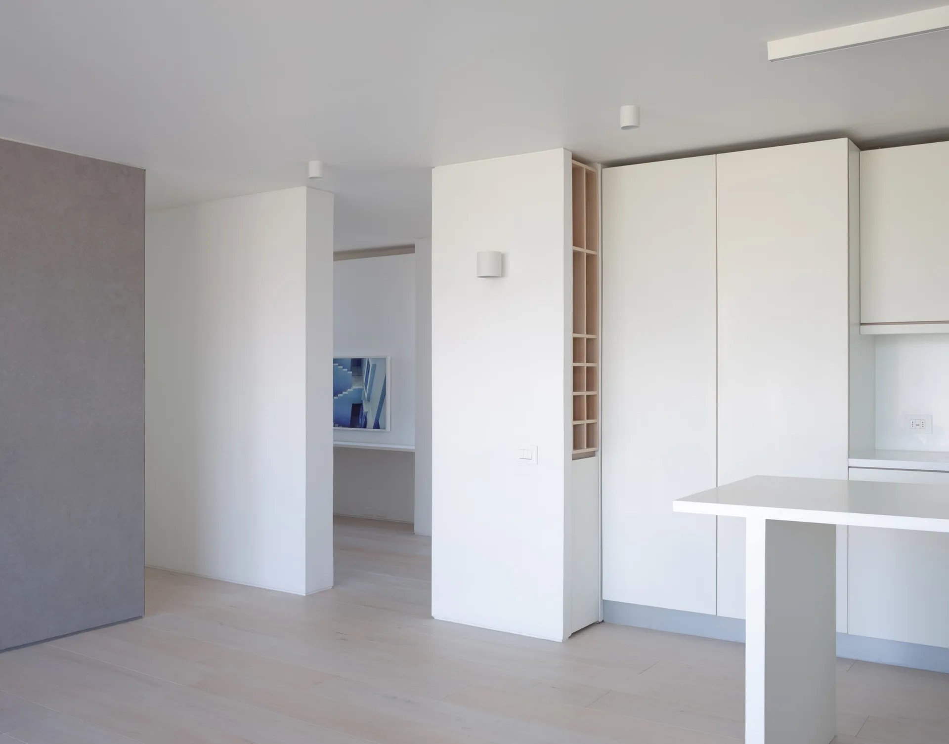 A minimalist white kitchen featuring seamless flat-front cabinetry, a vertical wood open shelving unit, a white island, and light wood floors.