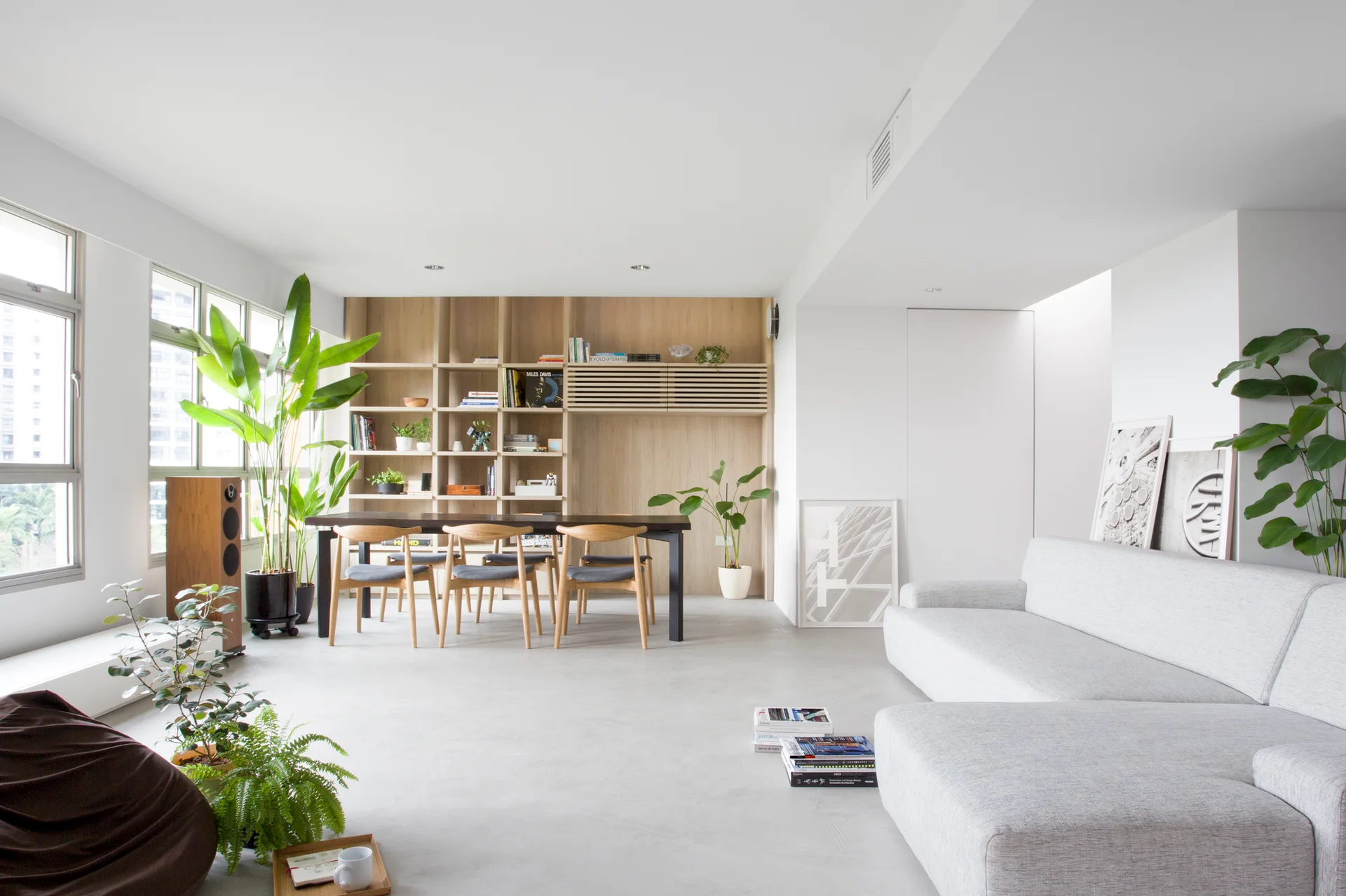 A bright open-concept living and dining space with a light grey sectional sofa, wooden dining table, and built-in light wood bookshelves.