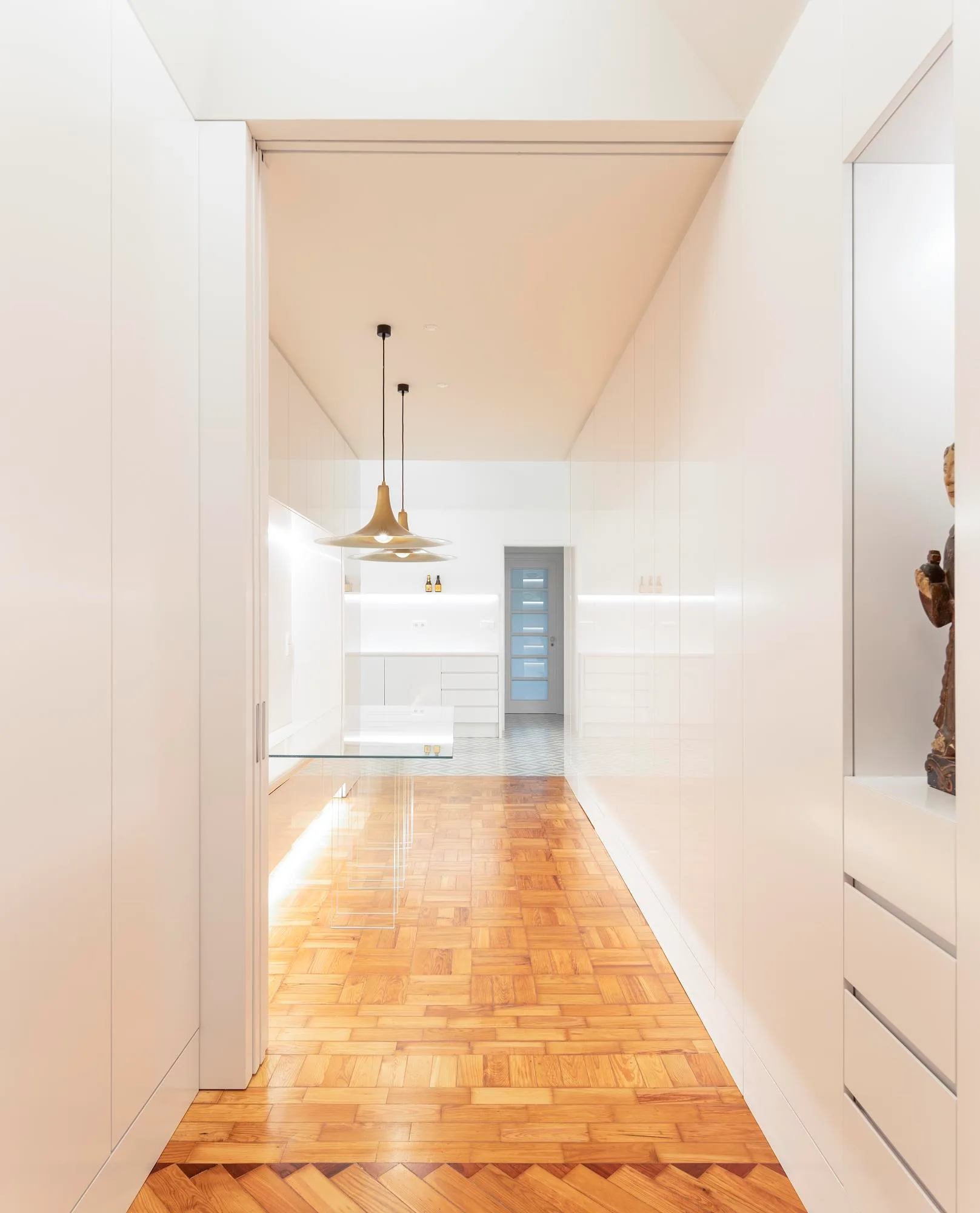 A minimalist white hallway featuring high-gloss built-in cabinets, warm parquet wood flooring, and brass pendant lights over a glass table.