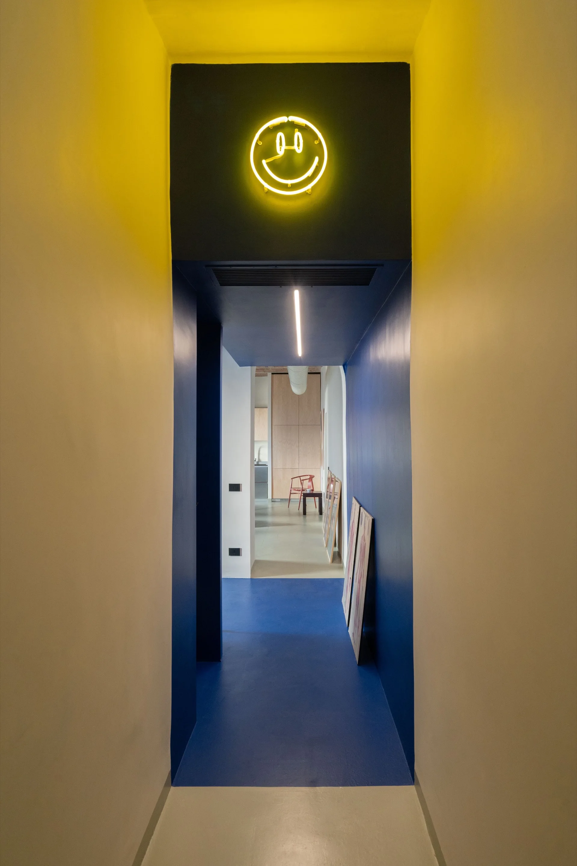A narrow hallway painted in dark blue features a glowing yellow smiley face neon sign above the doorway and framed art leaning on the floor.