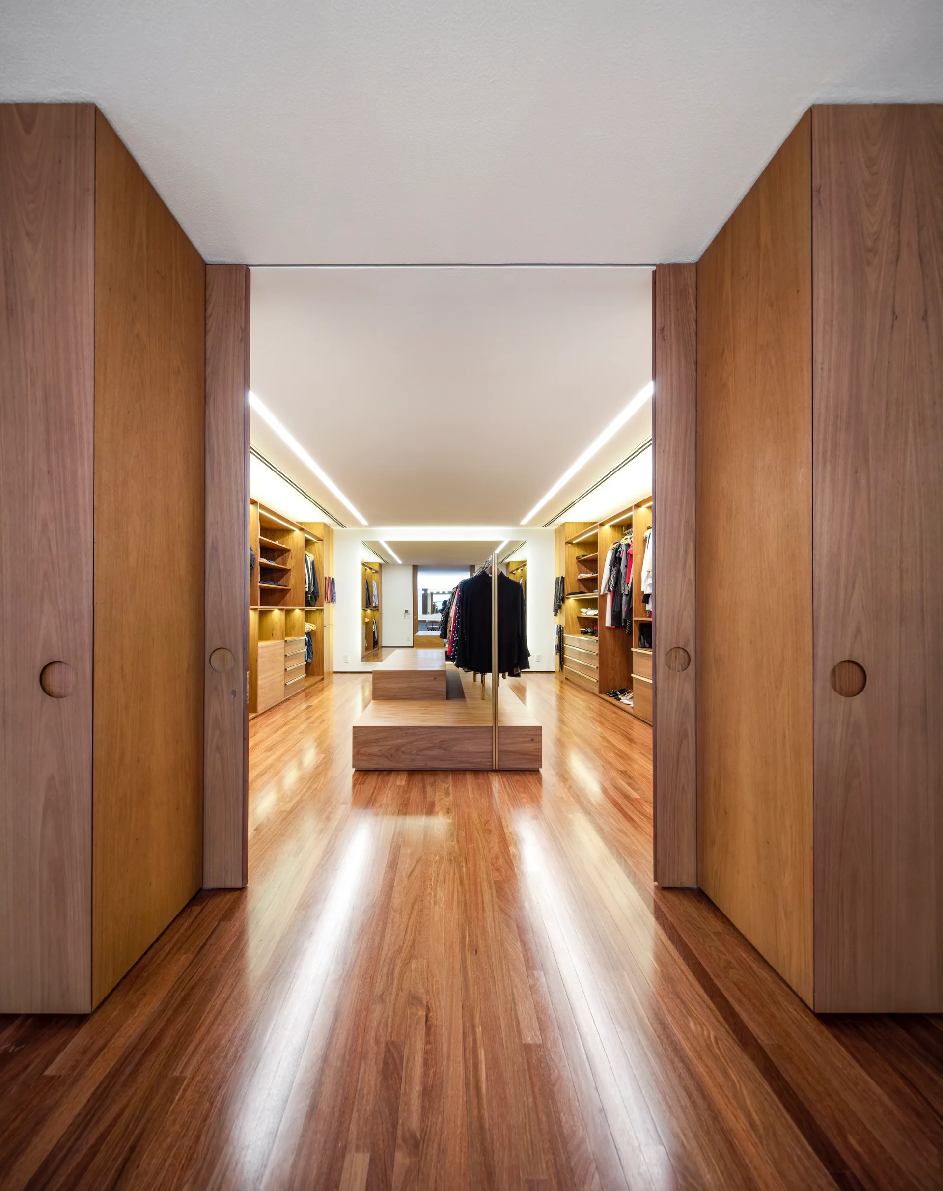 A spacious walk-in closet viewed through open wood sliding doors, featuring warm wood cabinetry, a polished floor, and a central tiered island.