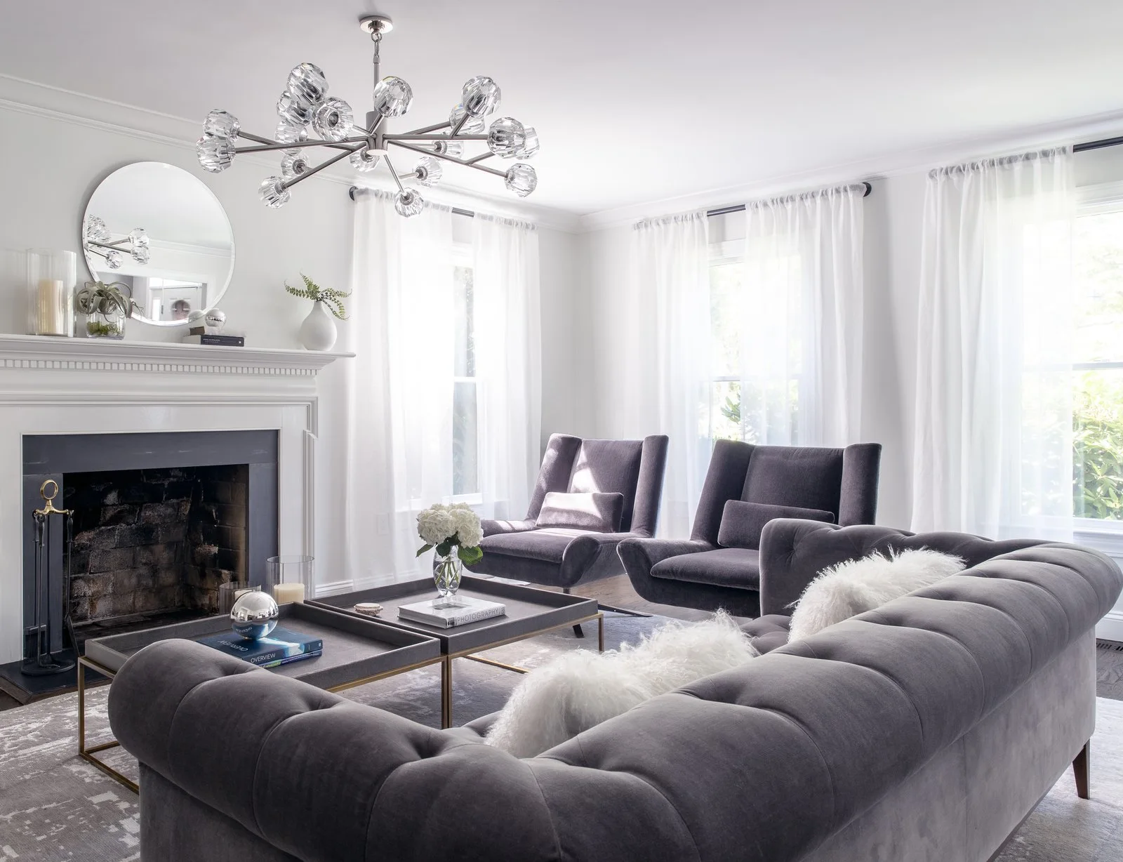 A living room with a grey tufted velvet sofa, matching armchairs, a fireplace with a round mirror, and a branching glass globe chandelier.