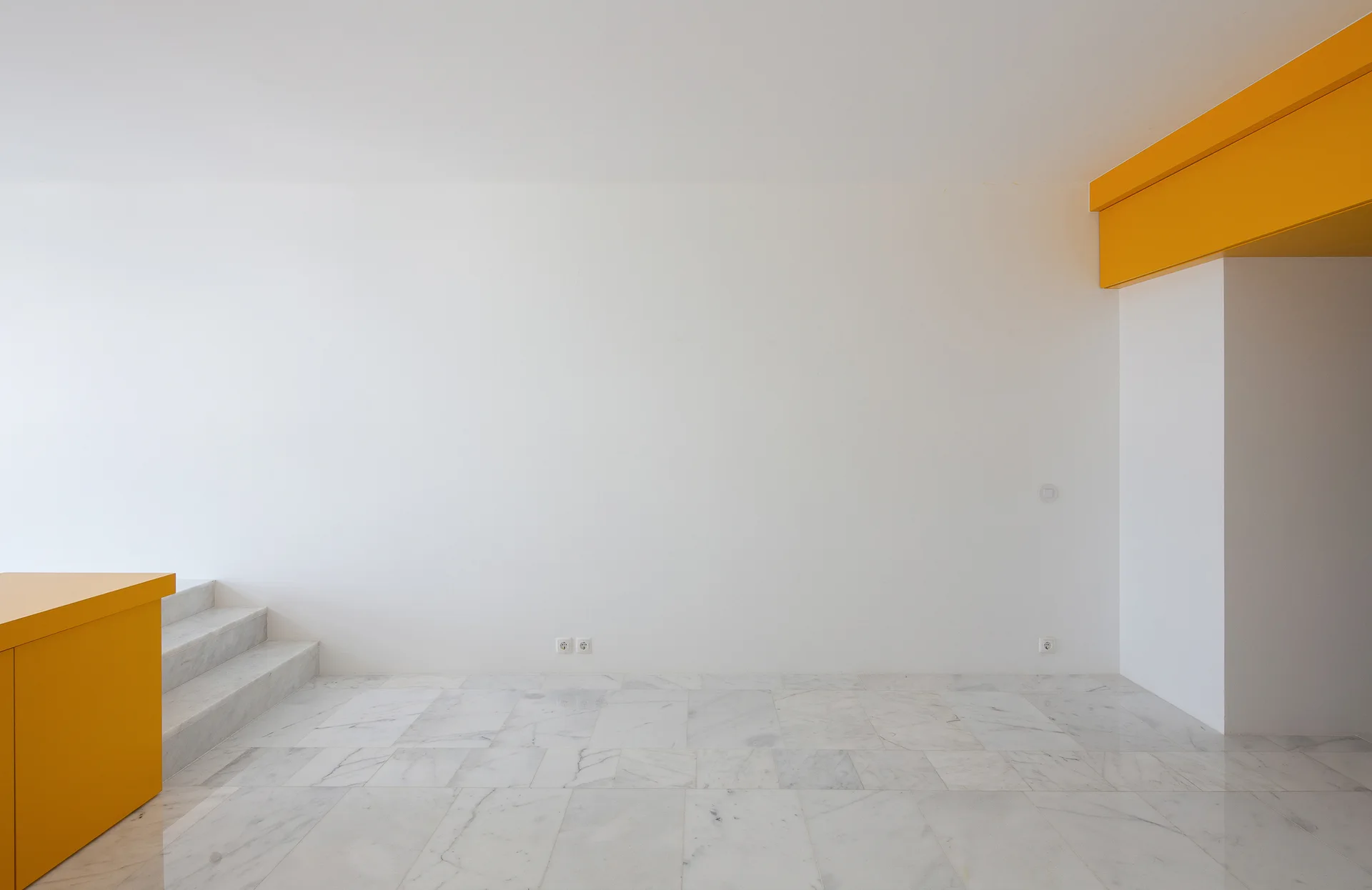 A minimalist room features polished white marble tile flooring, plain plaster walls, and bold yellow architectural details including a soffit and counter.