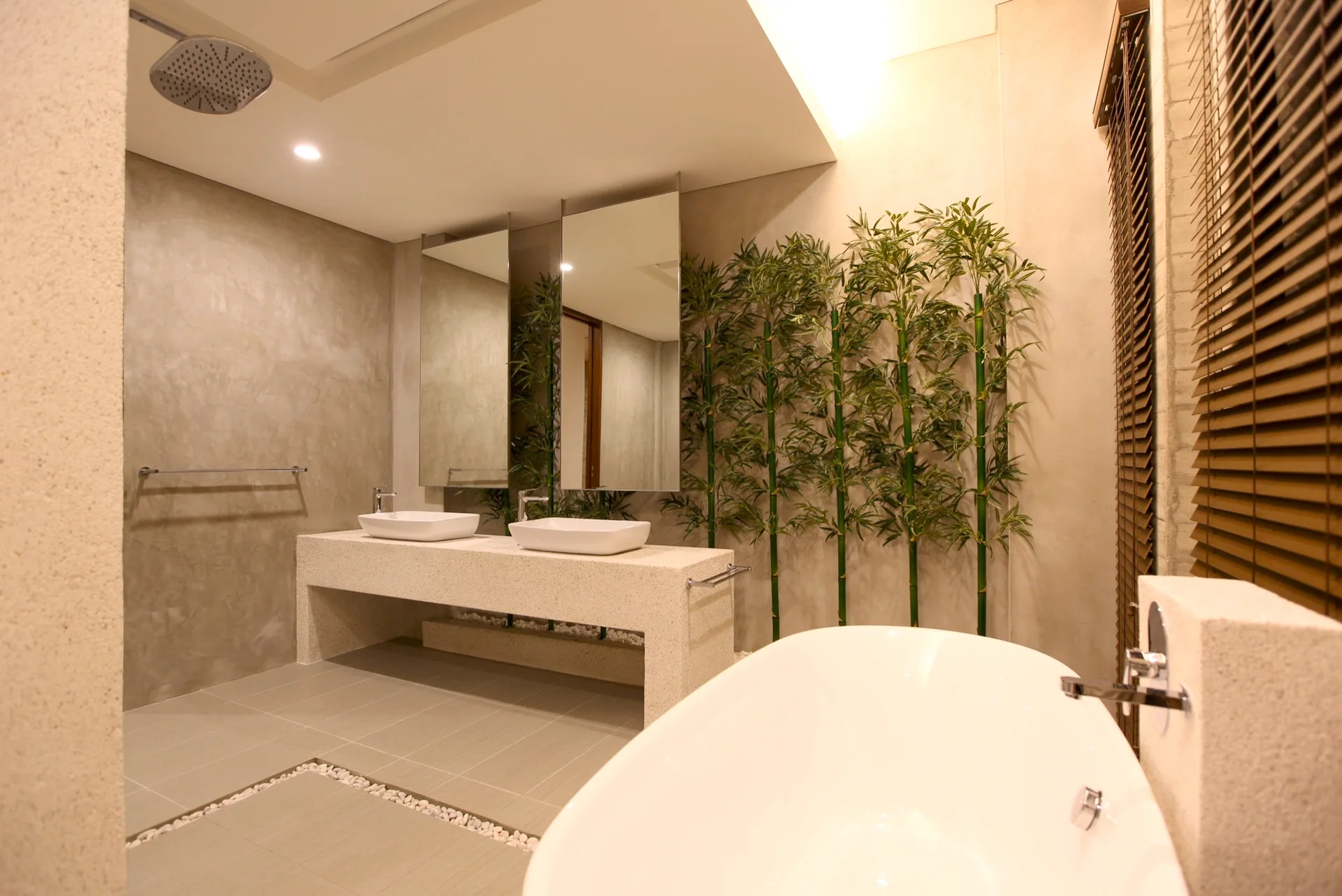 A modern bathroom featuring a white freestanding bathtub, a stone double vanity with vessel sinks, and green bamboo stalks against grey concrete walls.