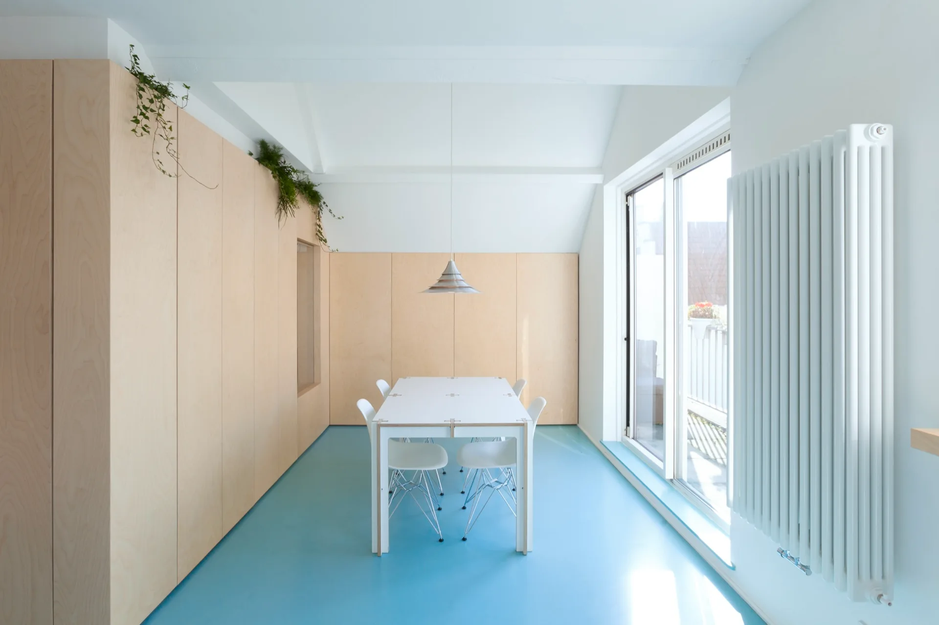 A minimalist dining room features a white table and chairs on a bright blue floor, set against a wall of light wood built-in cabinets.
