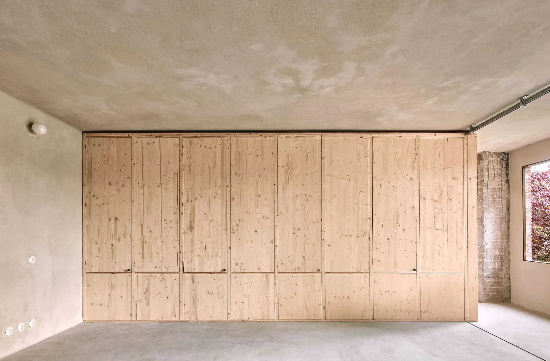 An expansive wall of floor-to-ceiling knotty wood cabinetry stands inside a minimalist room with raw concrete walls, ceiling, and floors.