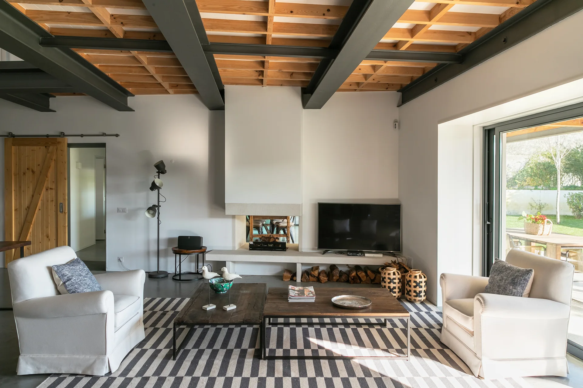 A living room featuring white armchairs, a black and white striped rug, low wooden coffee tables, and a ceiling with exposed wood and metal beams.