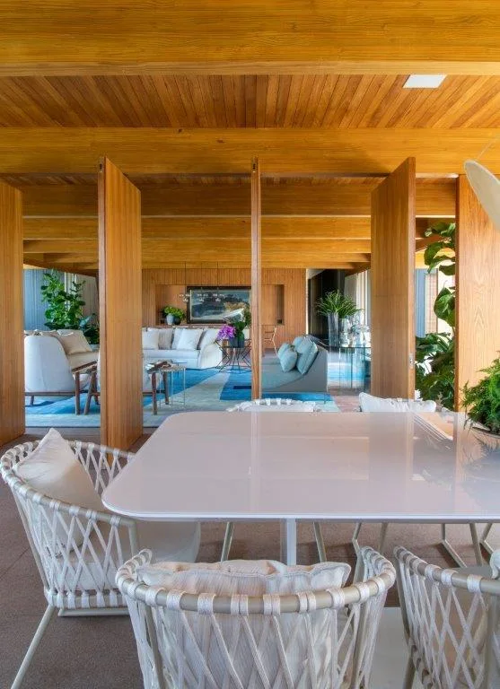 A glossy white dining table and woven chairs sit under a beamed wood ceiling, looking into a living room separated by vertical wood panels.