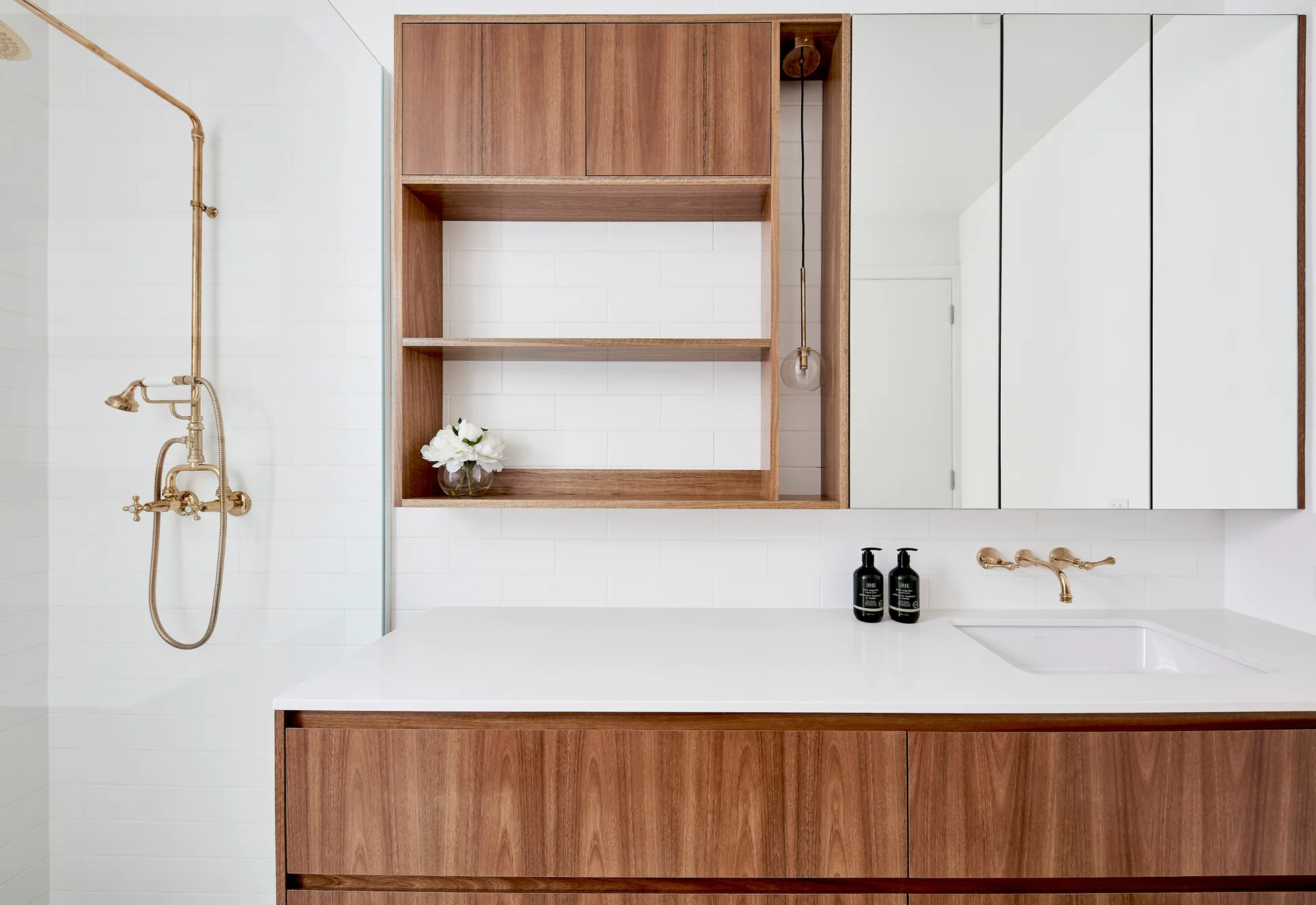 A modern bathroom features a wood vanity with a white countertop, gold wall-mounted faucet, mirrored medicine cabinet, and an exposed gold shower fixture.
