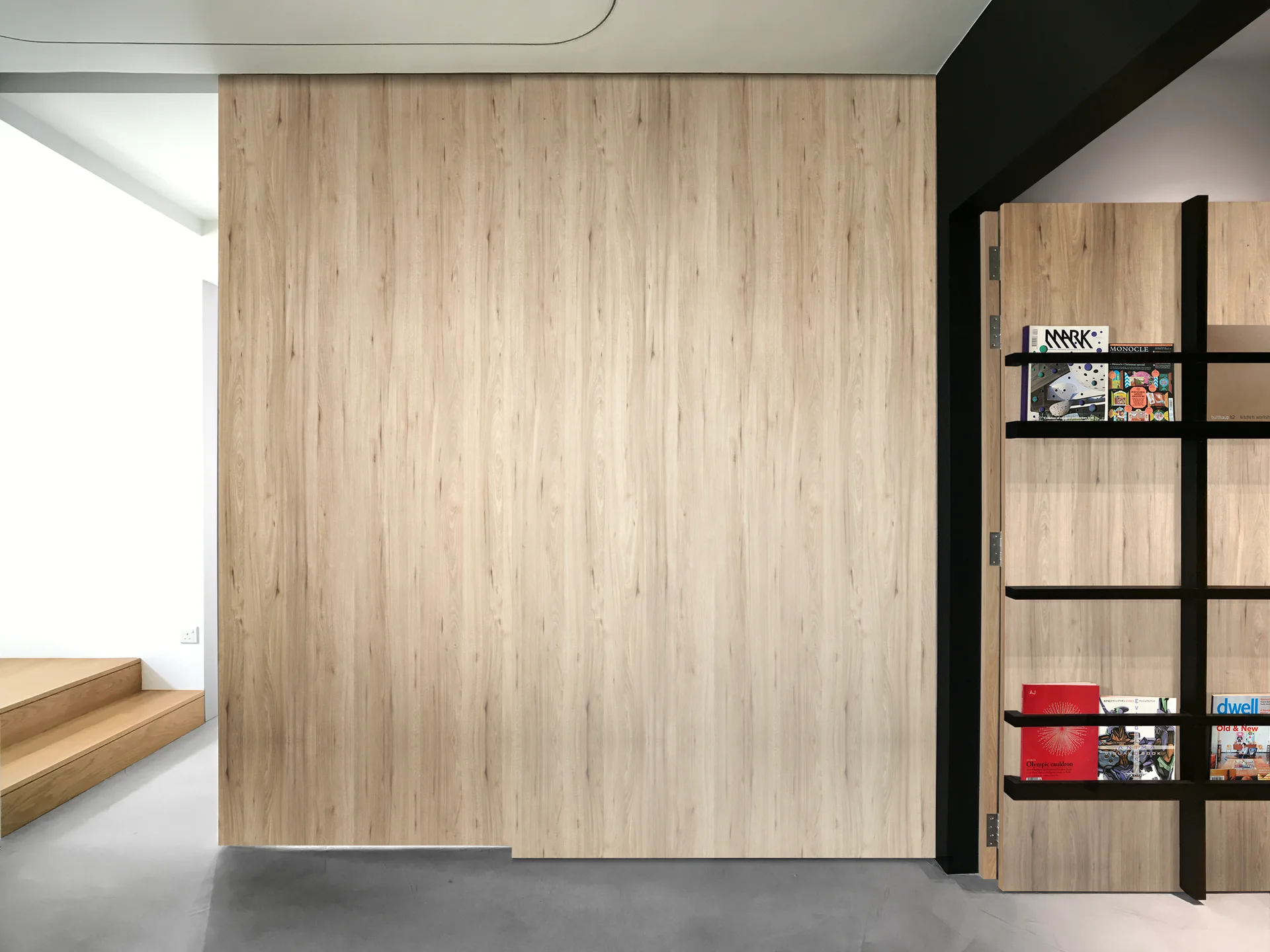 A modern hallway features a seamless light wood wall panel beside a black-framed vertical bookshelf displaying magazines on a grey concrete floor.
