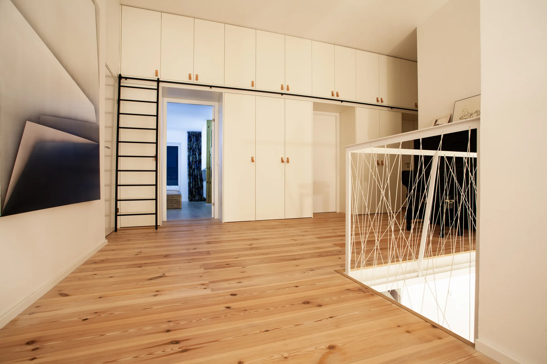 A minimalist hallway landing featuring a wall of white built-in cabinetry with a black sliding ladder, light wood flooring, and a white geometric railing.