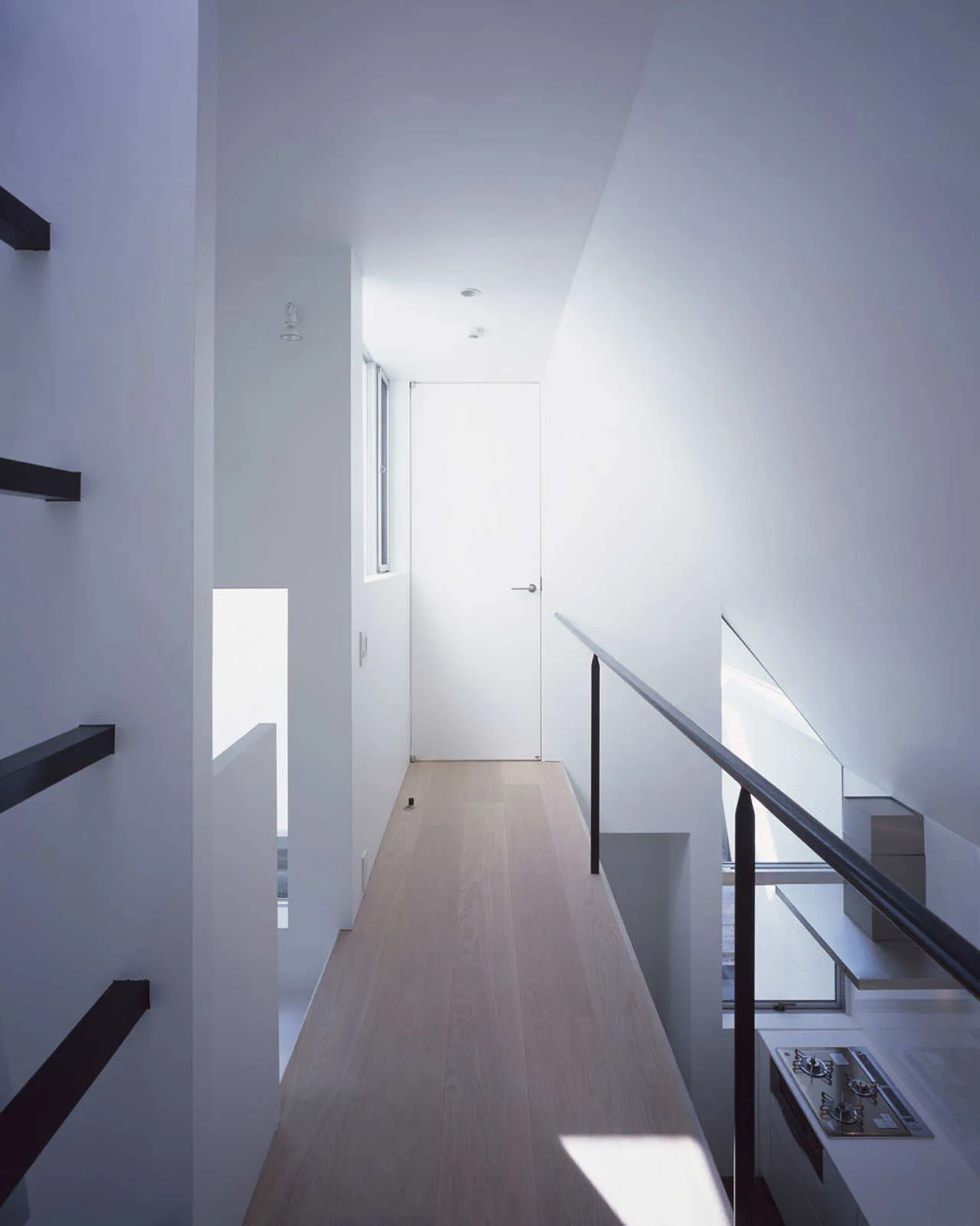 A narrow minimalist hallway with light wood flooring, stark white walls, and a slim black metal railing overlooking the kitchen below.