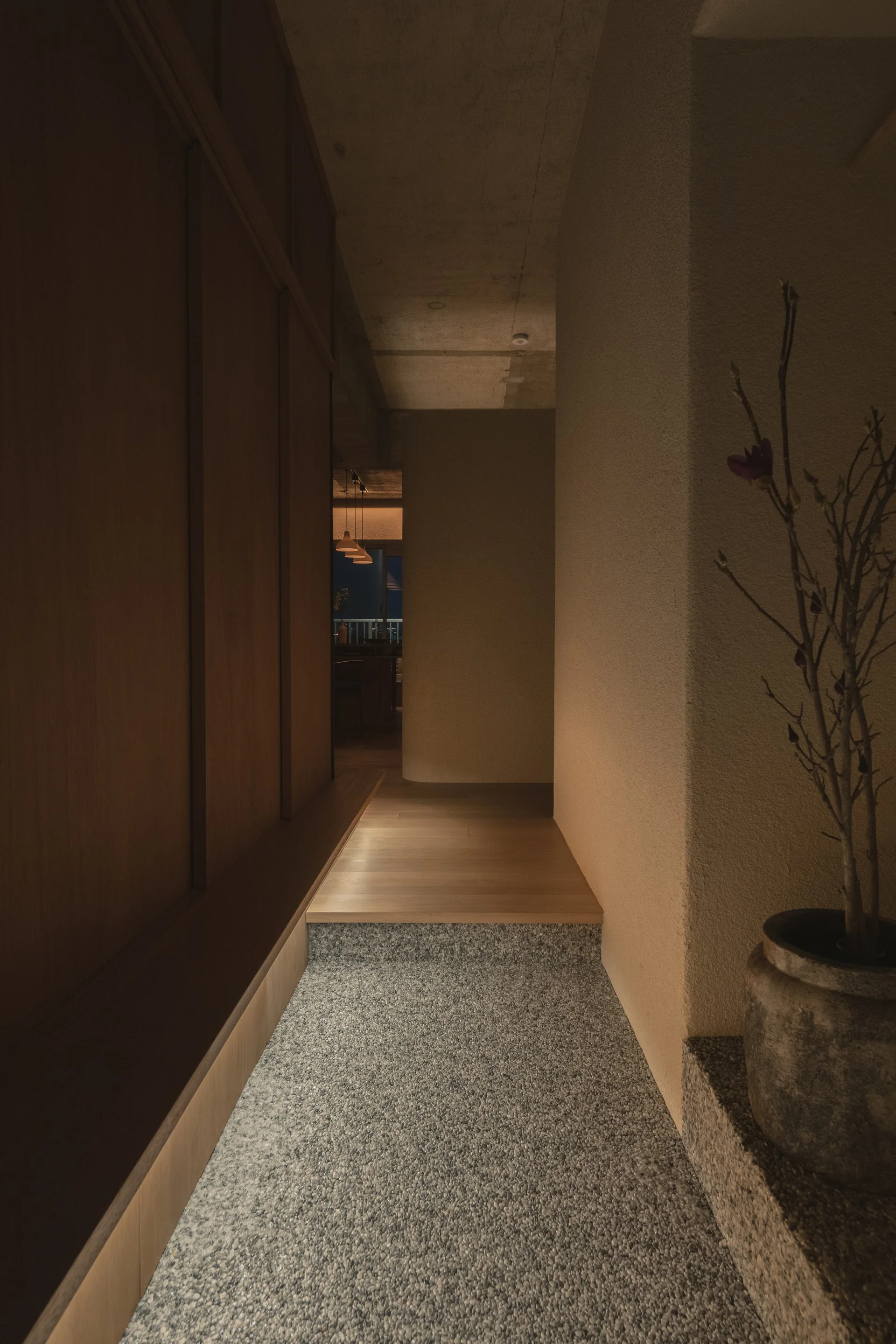 A minimalist entryway features a textured pebbled stone floor transitioning to a raised wood floor step alongside full-height wood paneling.