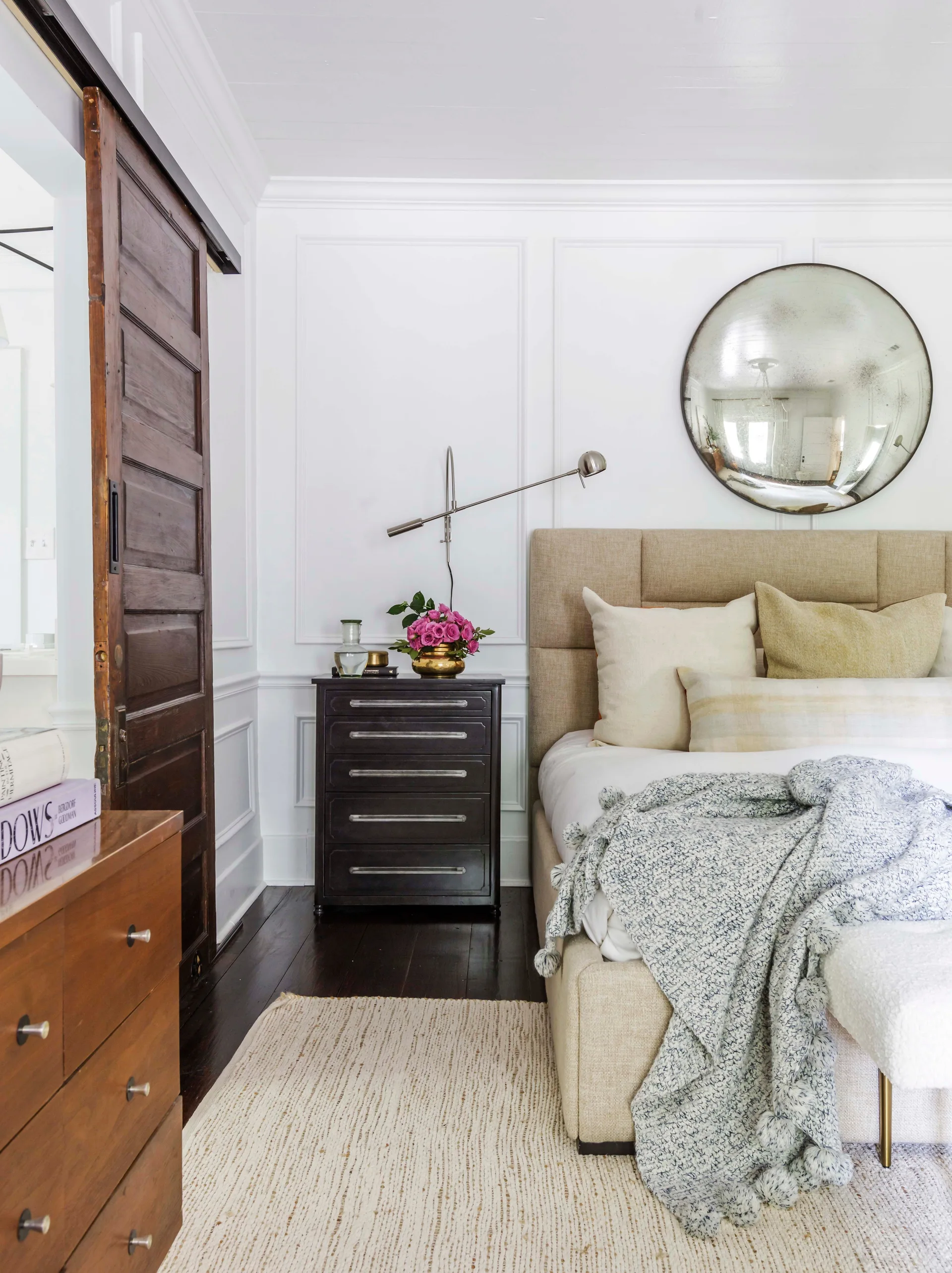 An interior view of a bedroom featuring an upholstered bed, a dark wood sliding barn door, and white paneled walls with a round mirror.