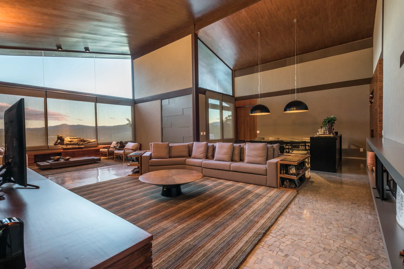 A spacious living room featuring a large grey sectional sofa, a striped area rug, a round wooden coffee table, and a vaulted wood-paneled ceiling.