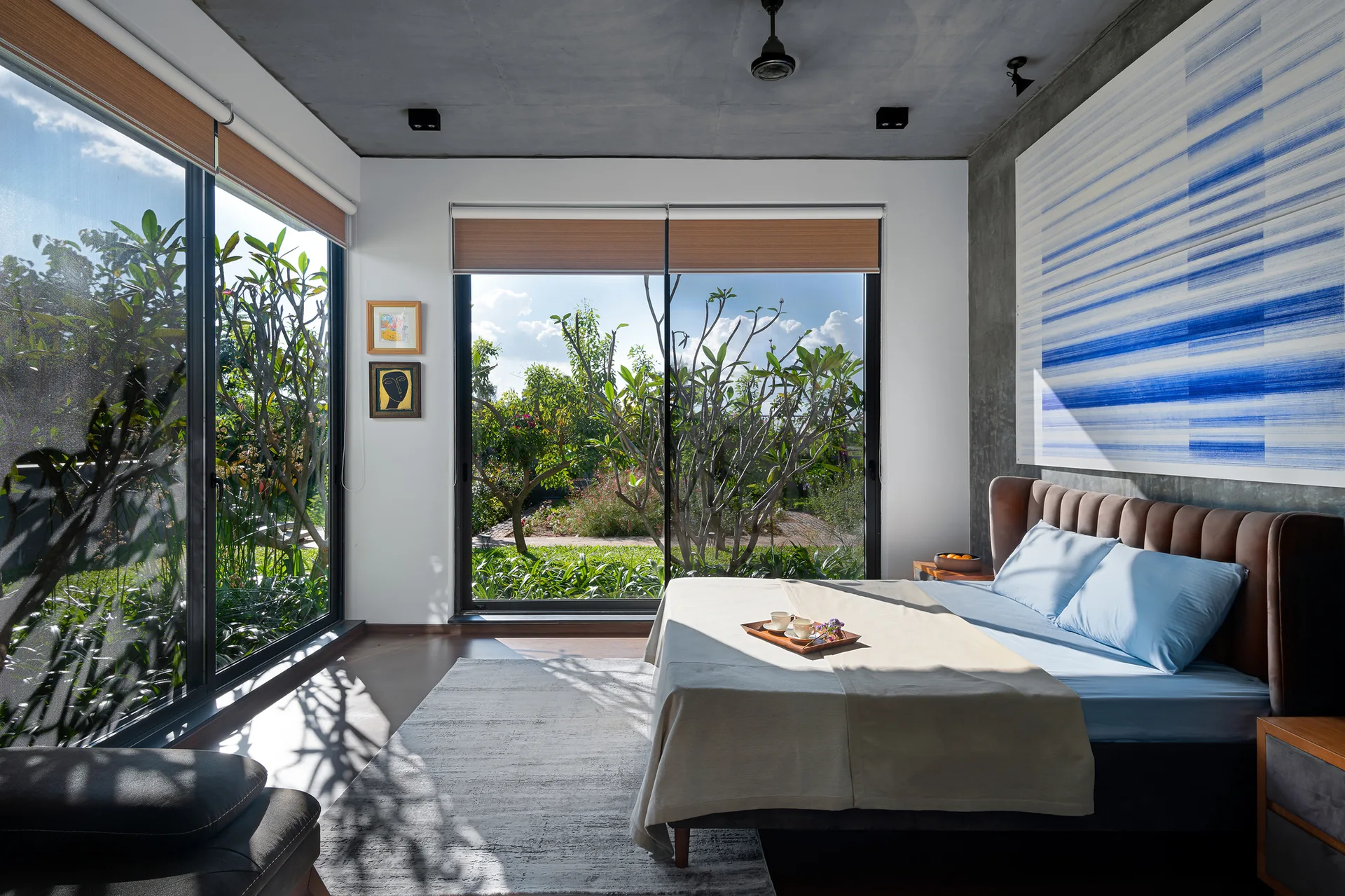 A modern bedroom features a large bed with a brown headboard, concrete ceilings, and floor-to-ceiling windows overlooking a lush garden.