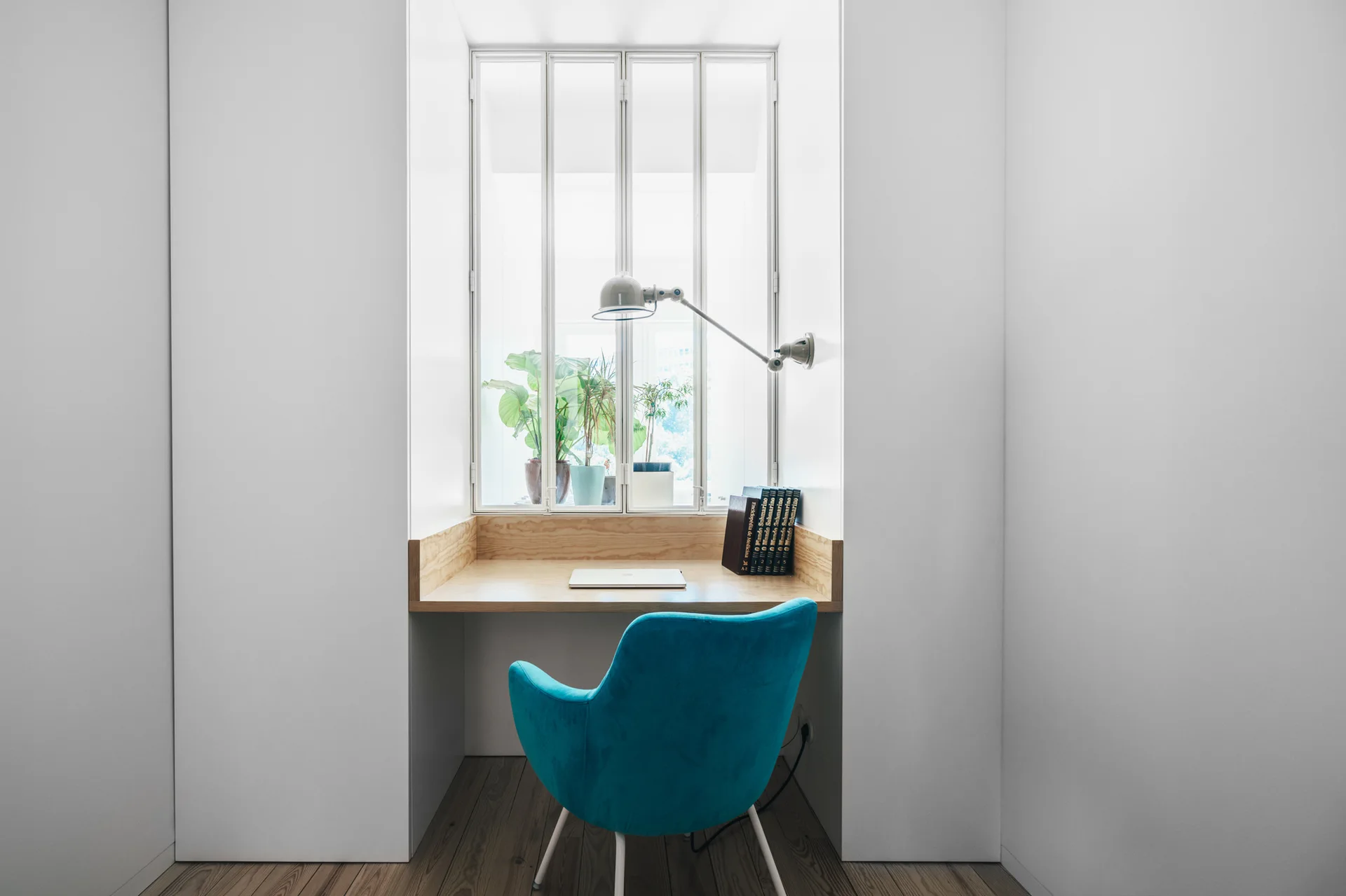 A built-in wooden desk nestled in a white wall alcove beneath a window, paired with a bright blue upholstered chair.