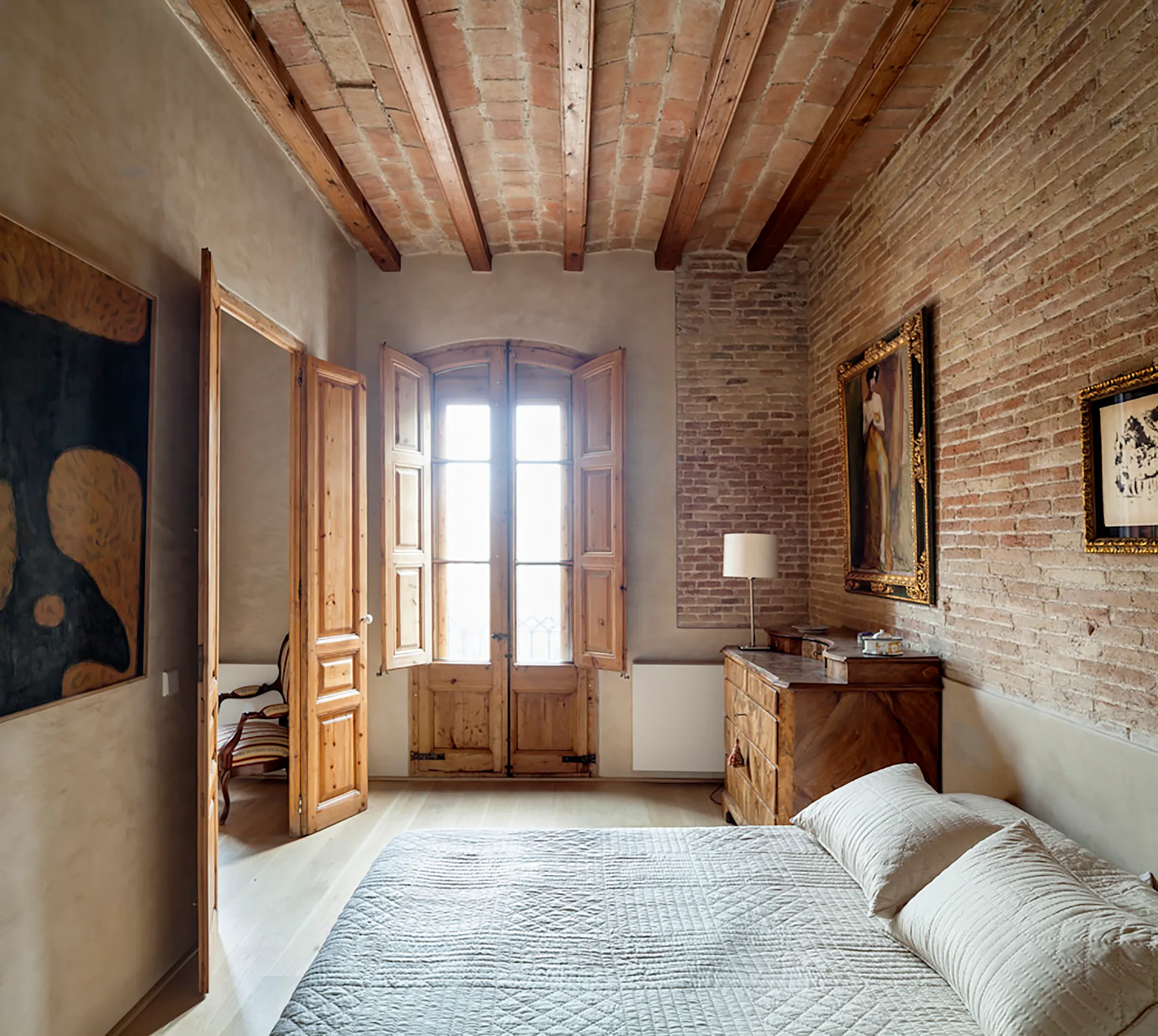 A rustic bedroom featuring exposed brick walls, a vaulted brick and wood-beam ceiling, a wooden chest of drawers, and an arched window with shutters.