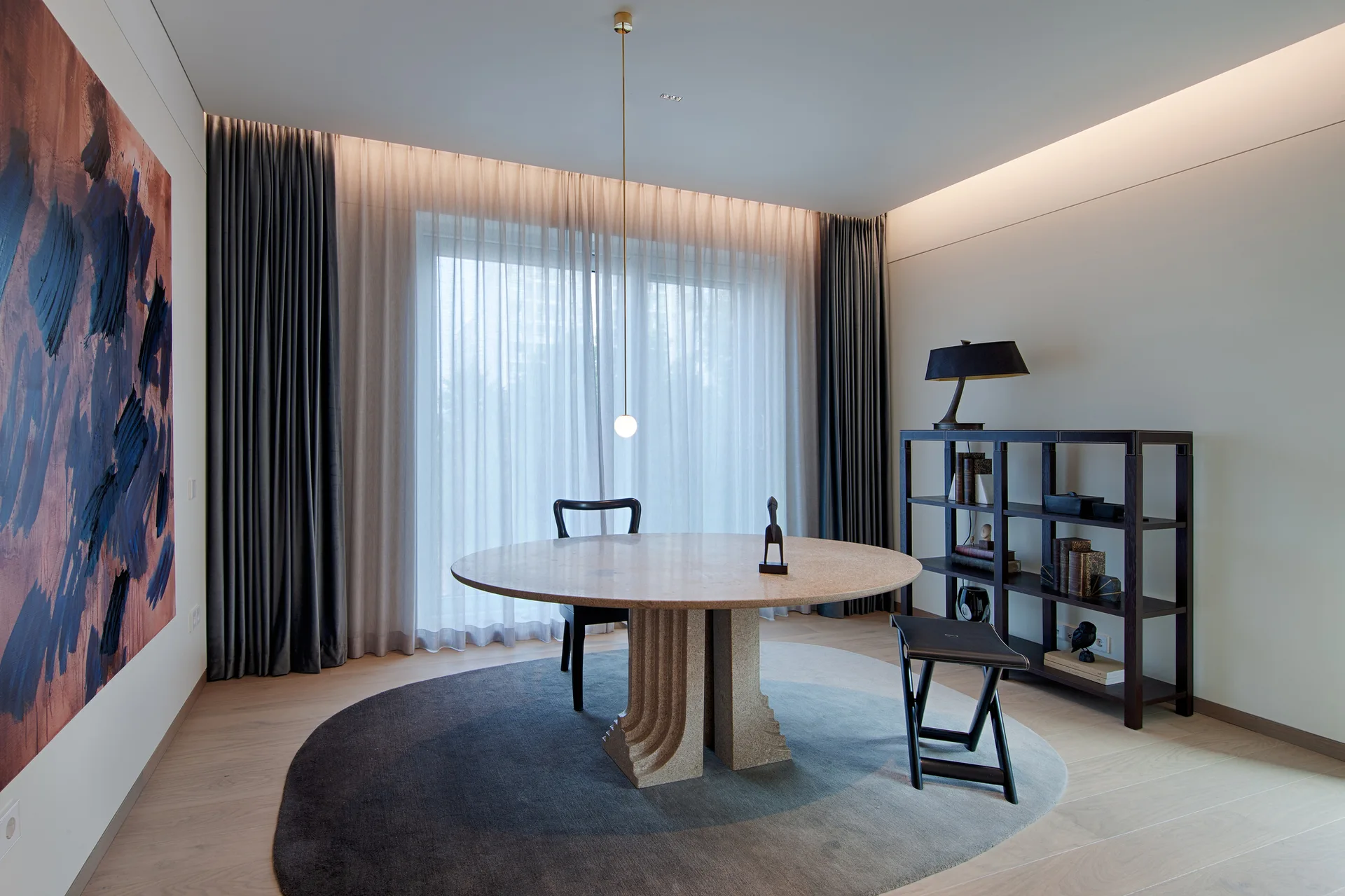 A modern dining room features a round stone table on a circular gradient rug next to a black open bookshelf and sheer grey drapery.