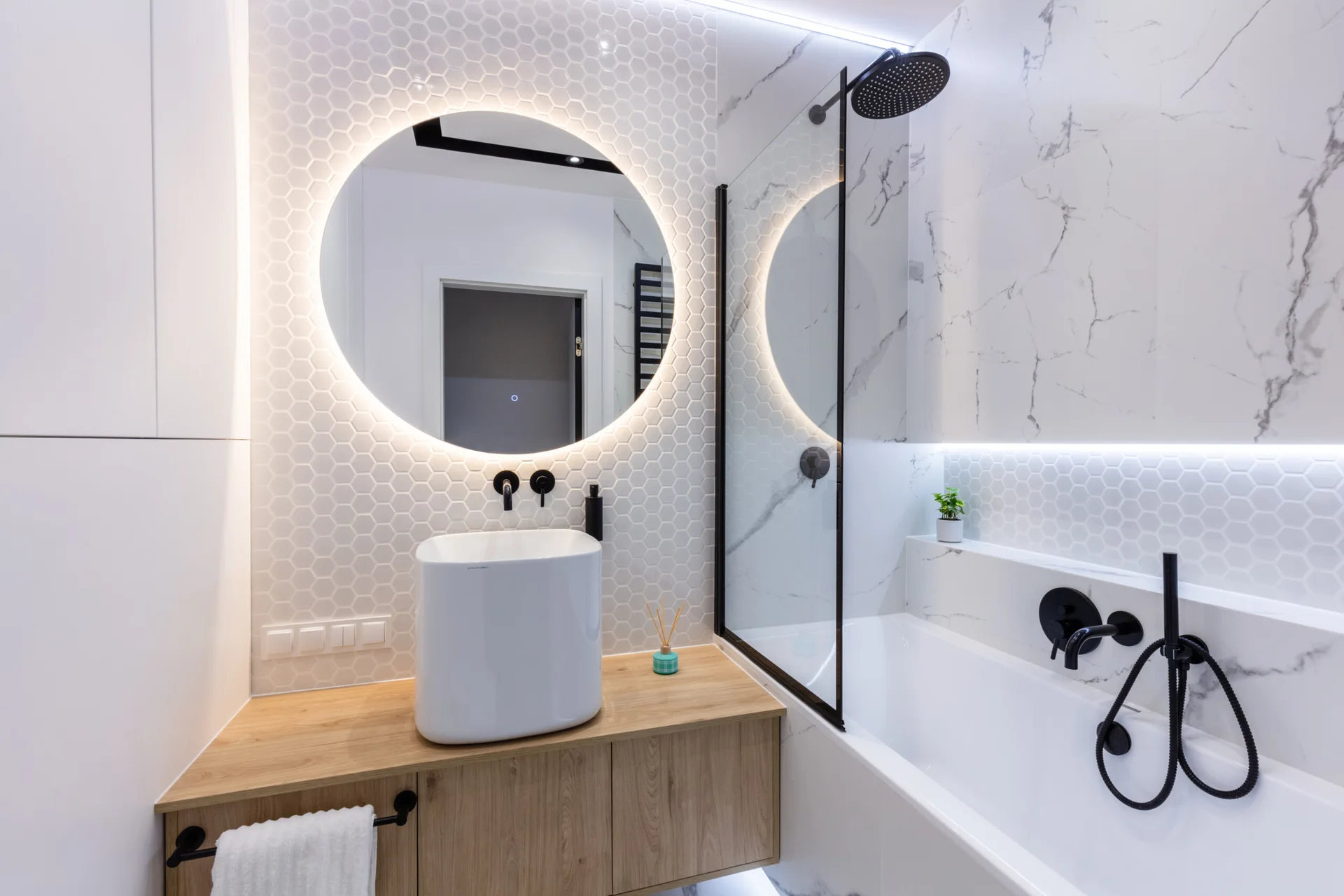 A modern bathroom features a round backlit mirror over a wood vanity with a white vessel sink next to a bathtub with matte black fixtures.