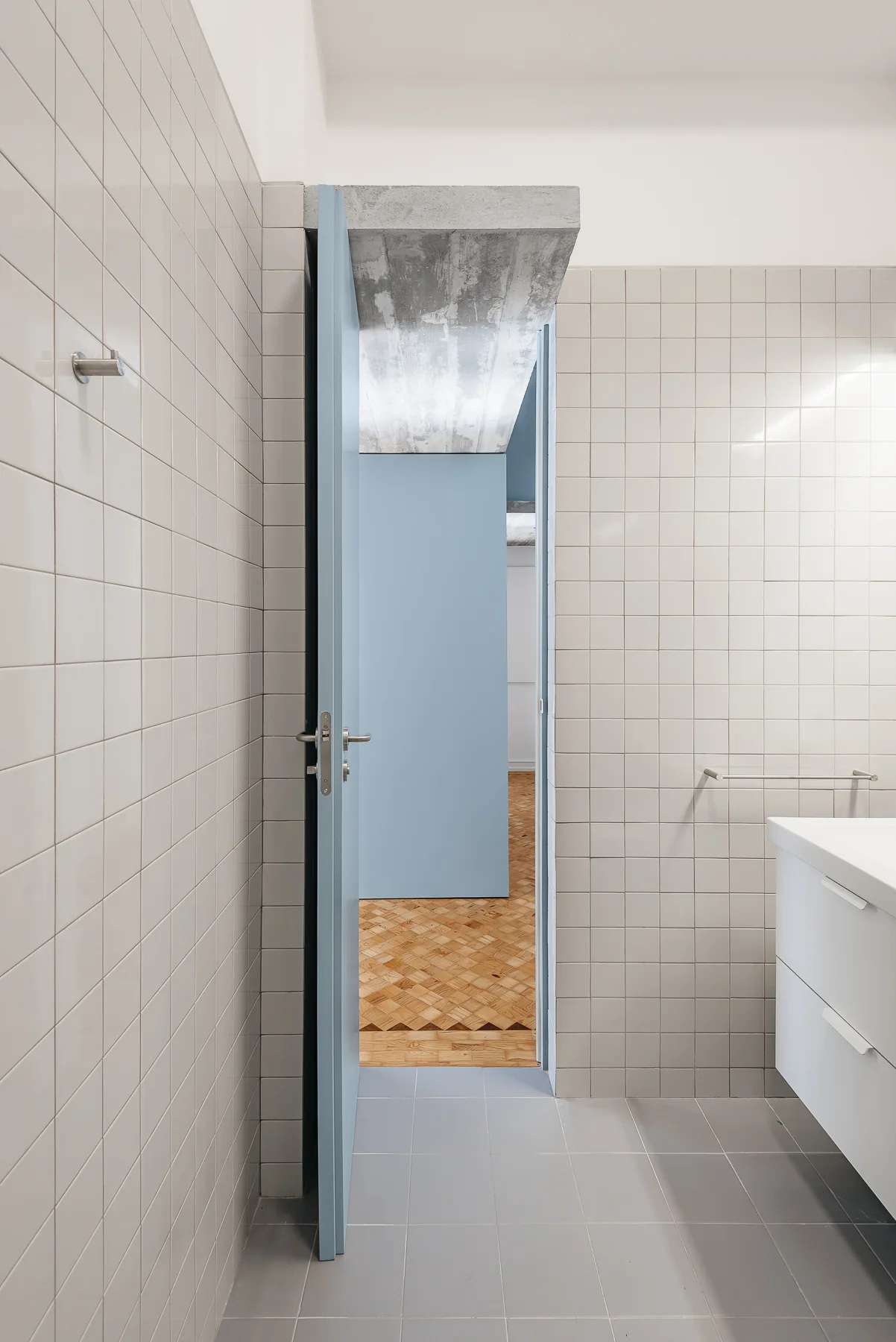 A modern bathroom with white square wall tiles and a light blue open door leading to an adjacent room with parquet wood flooring.