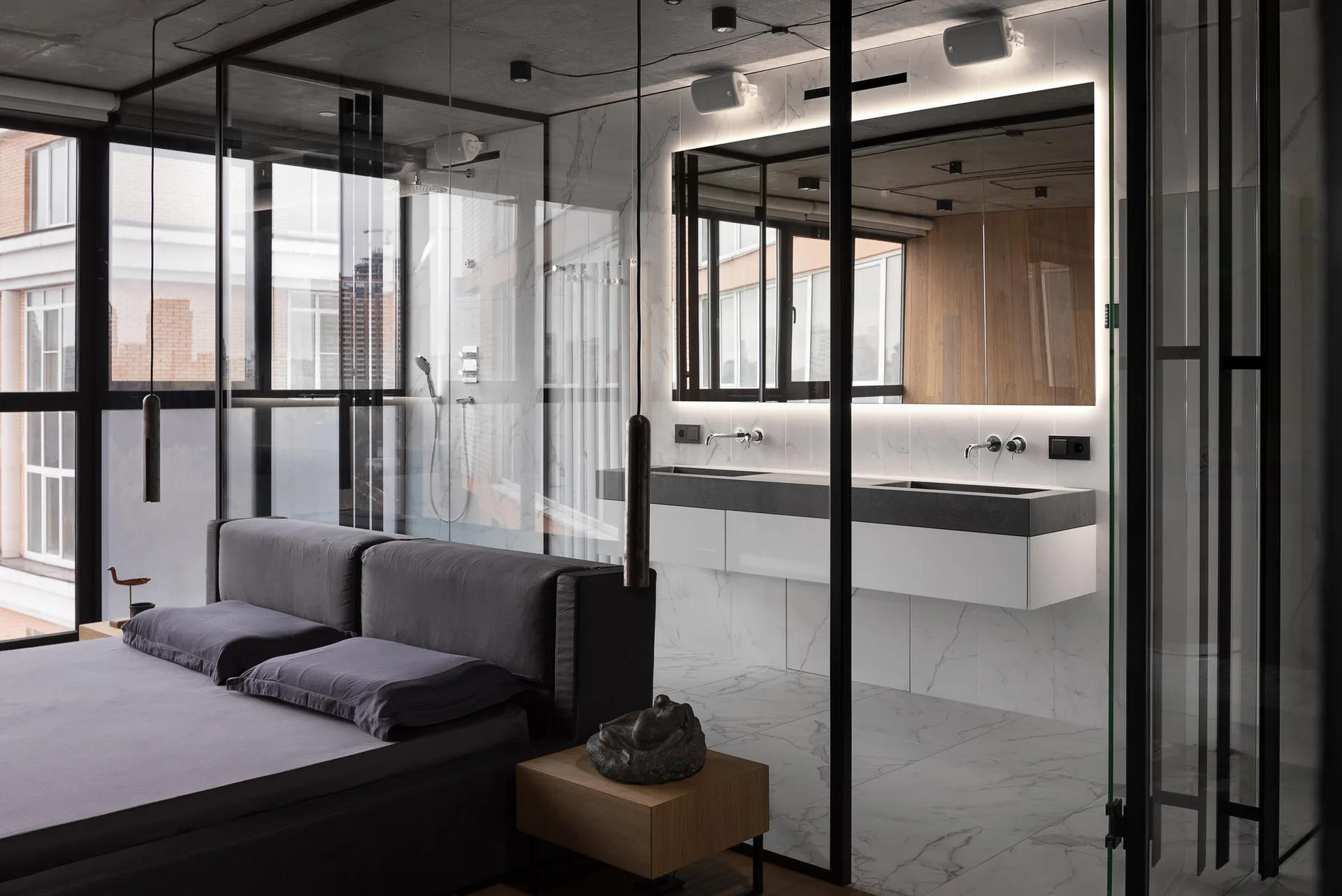 A modern bedroom features a grey platform bed next to a glass-partitioned ensuite bathroom with a floating white vanity.