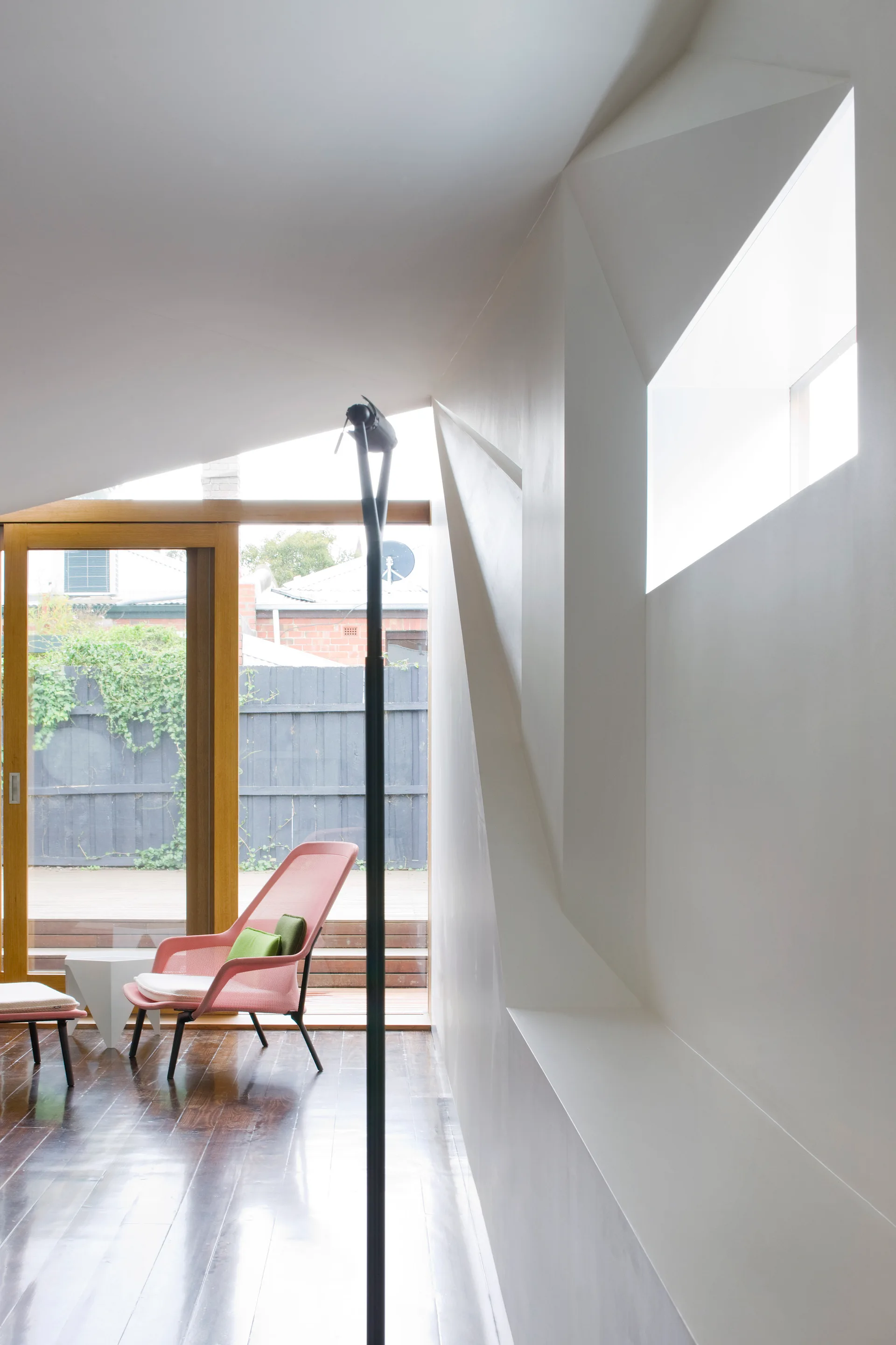 A pink mesh lounge chair sits near a wood-framed glass door in a white room with dark wood floors and an angular sloped ceiling with a skylight.