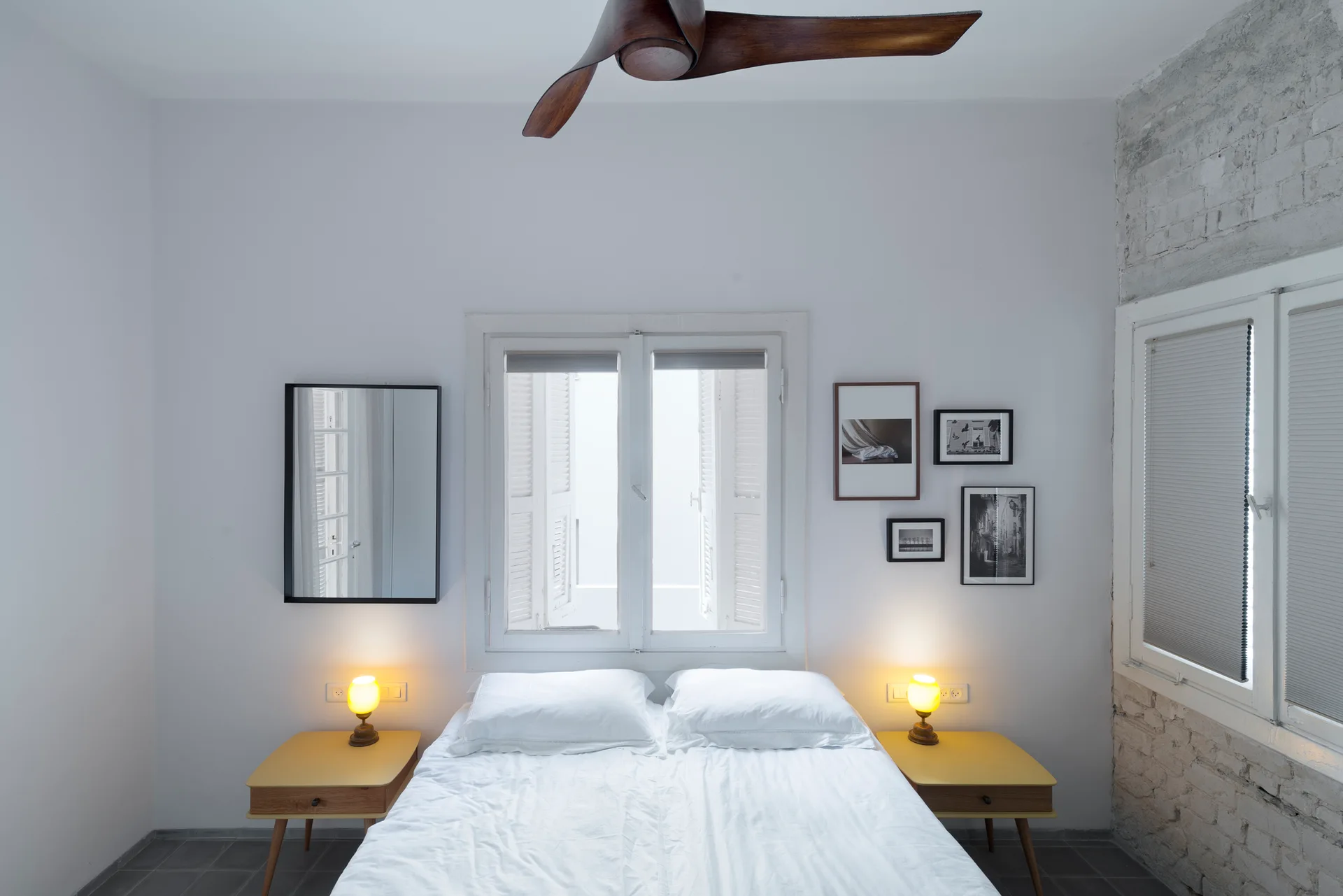 A minimalist white bedroom features a bed centered under a shuttered window, flanked by two yellow nightstands with glowing orb lamps.