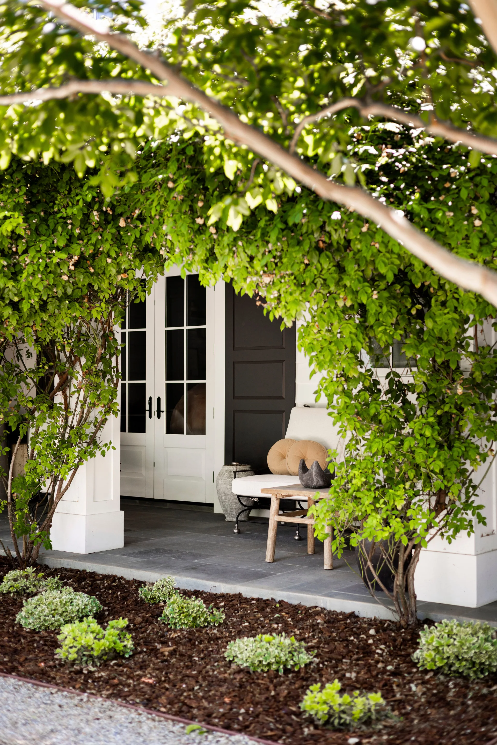 A covered stone porch features a white upholstered armchair, a rustic wooden side table, and white French doors framed by overgrown green trees.