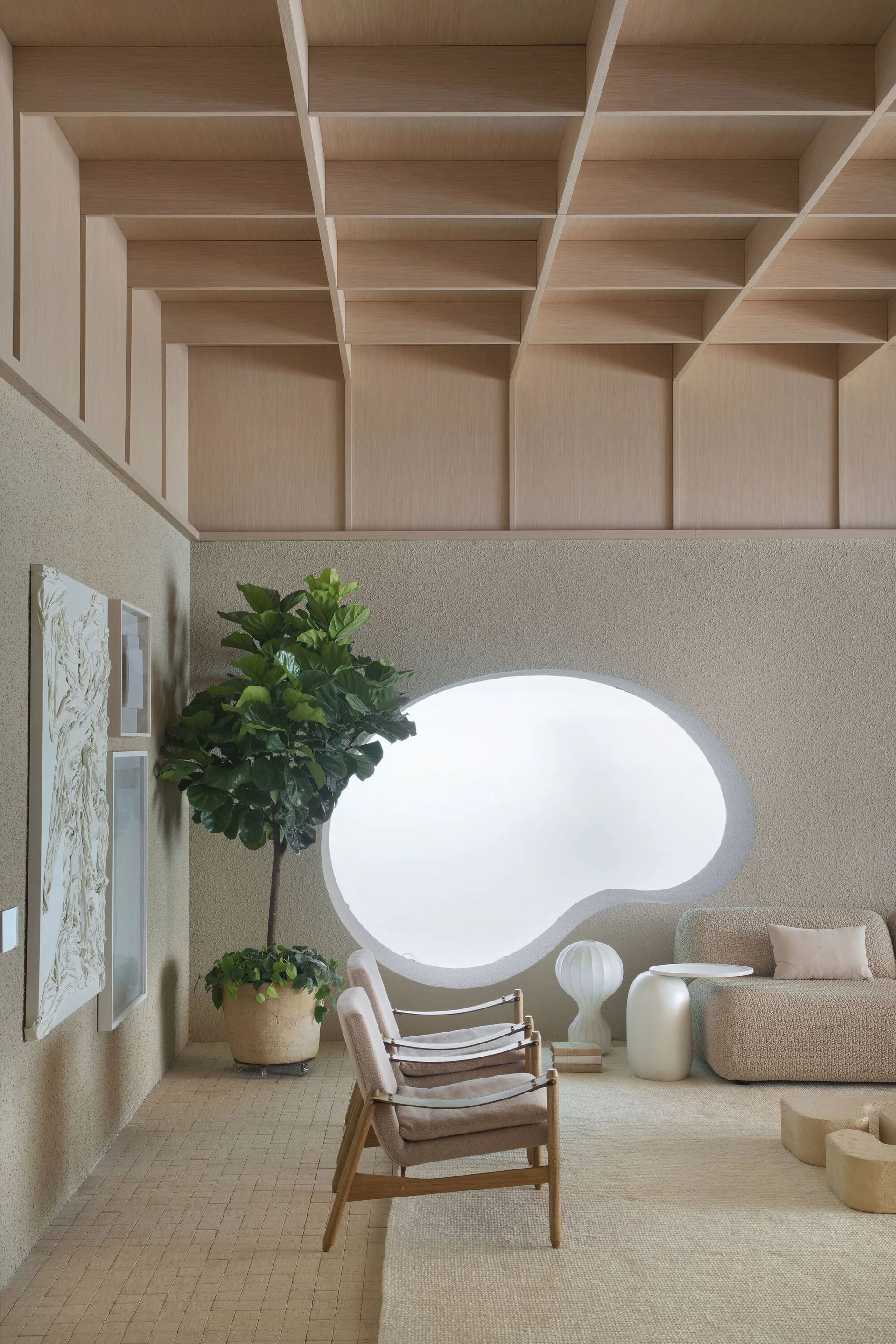 A minimalist living room features a large organic-shaped window, a geometric coffered wood ceiling, a textured beige sofa, and wood-framed lounge chairs.