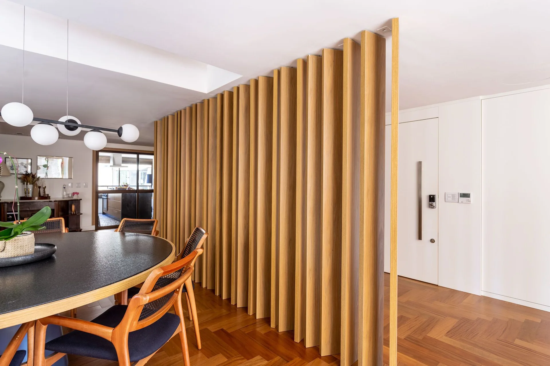 A modern dining area featuring a round black table, cane chairs, herringbone wood flooring, and a large vertical wood slat partition screen.