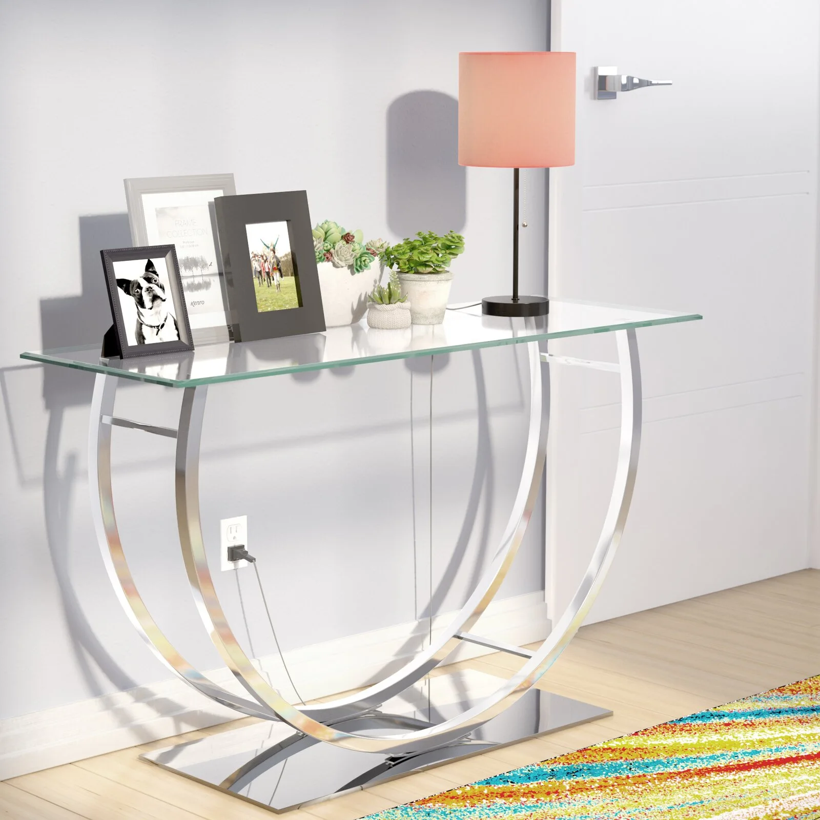 A modern glass console table with a polished chrome crescent base is decorated with picture frames, potted succulents, and a coral-shaded lamp.