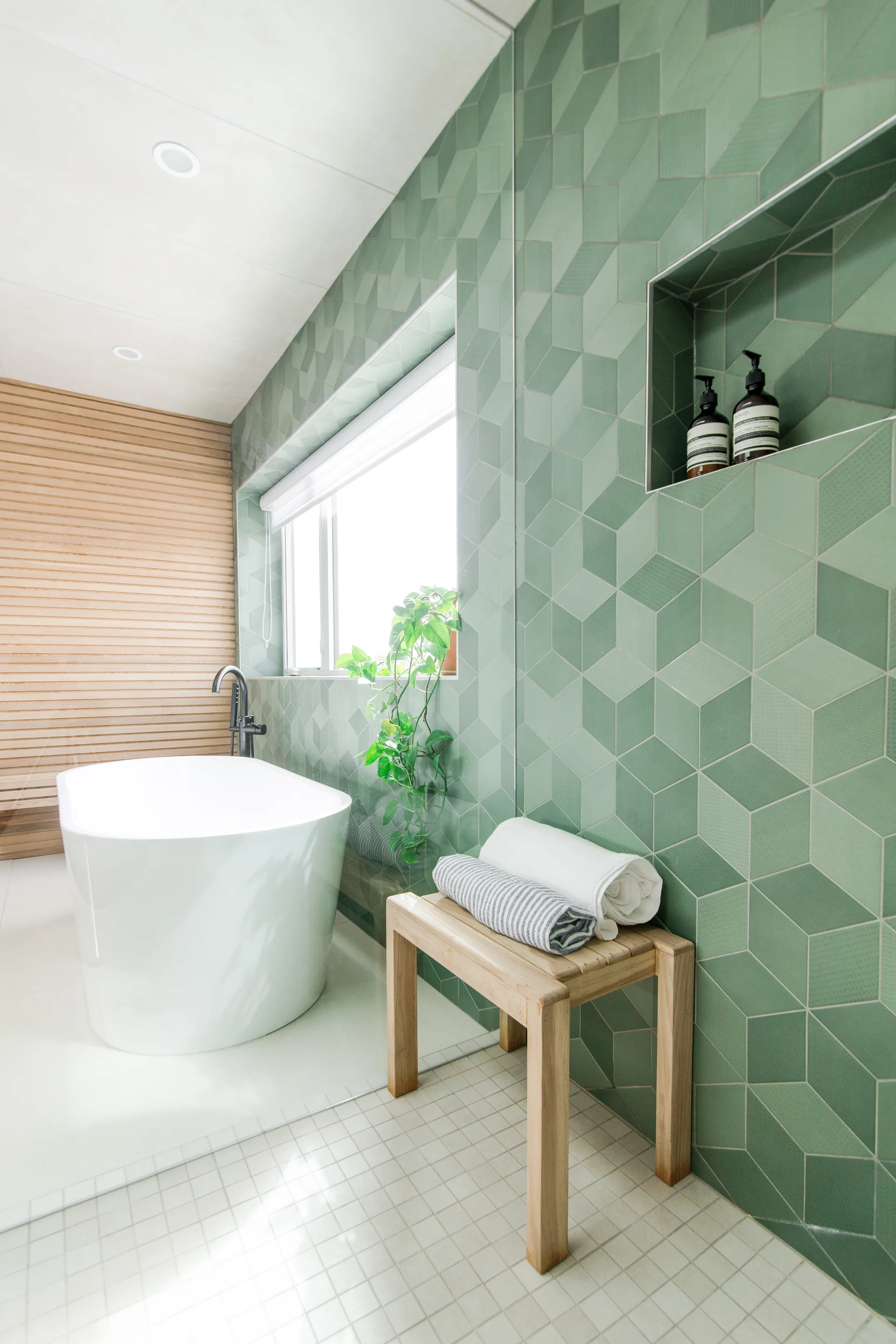 A modern bathroom with a white freestanding bathtub next to a sage green geometric tiled wall, a wooden stool, and a recessed wall niche.
