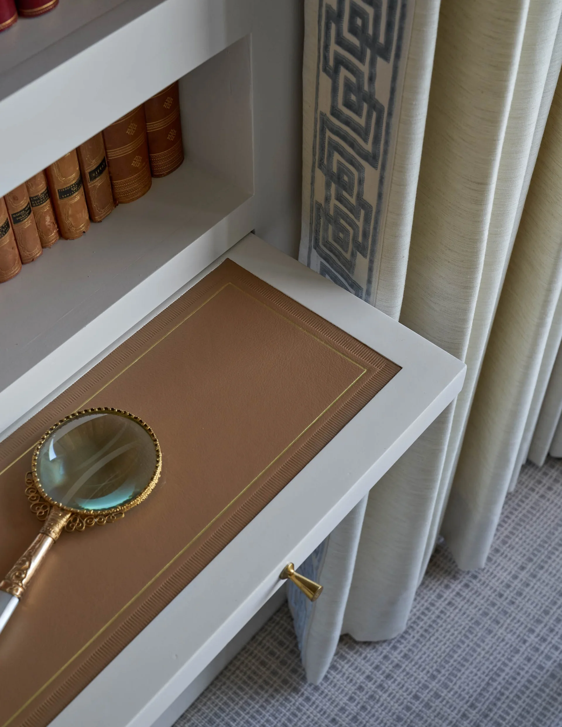 A close-up of a white built-in desk with a tan leather inset, holding an ornate magnifying glass next to cream drapes with Greek key trim.