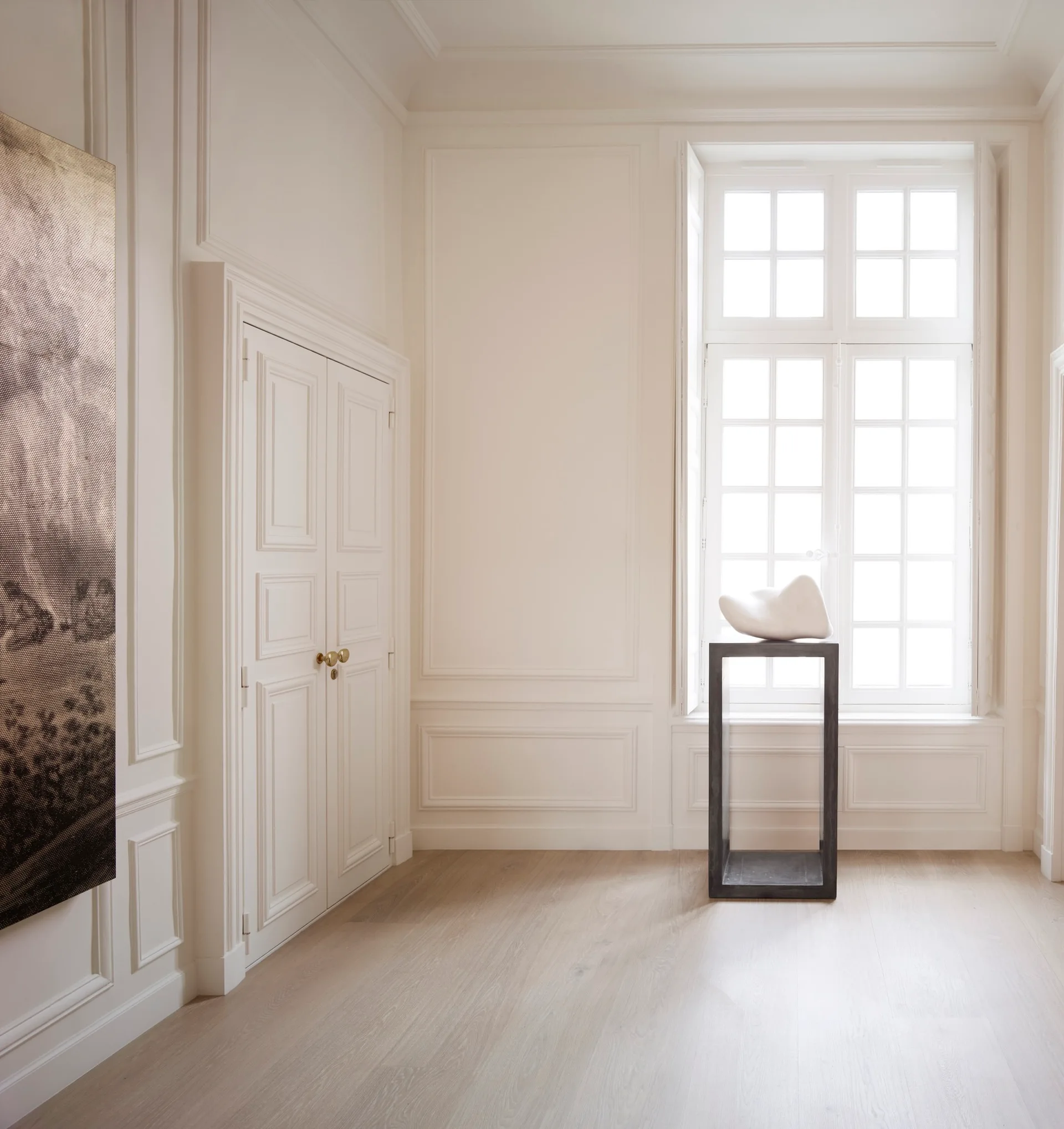 An off-white room features neoclassical wall moulding, double doors, and a black pedestal holding an abstract sculpture in front of a window.