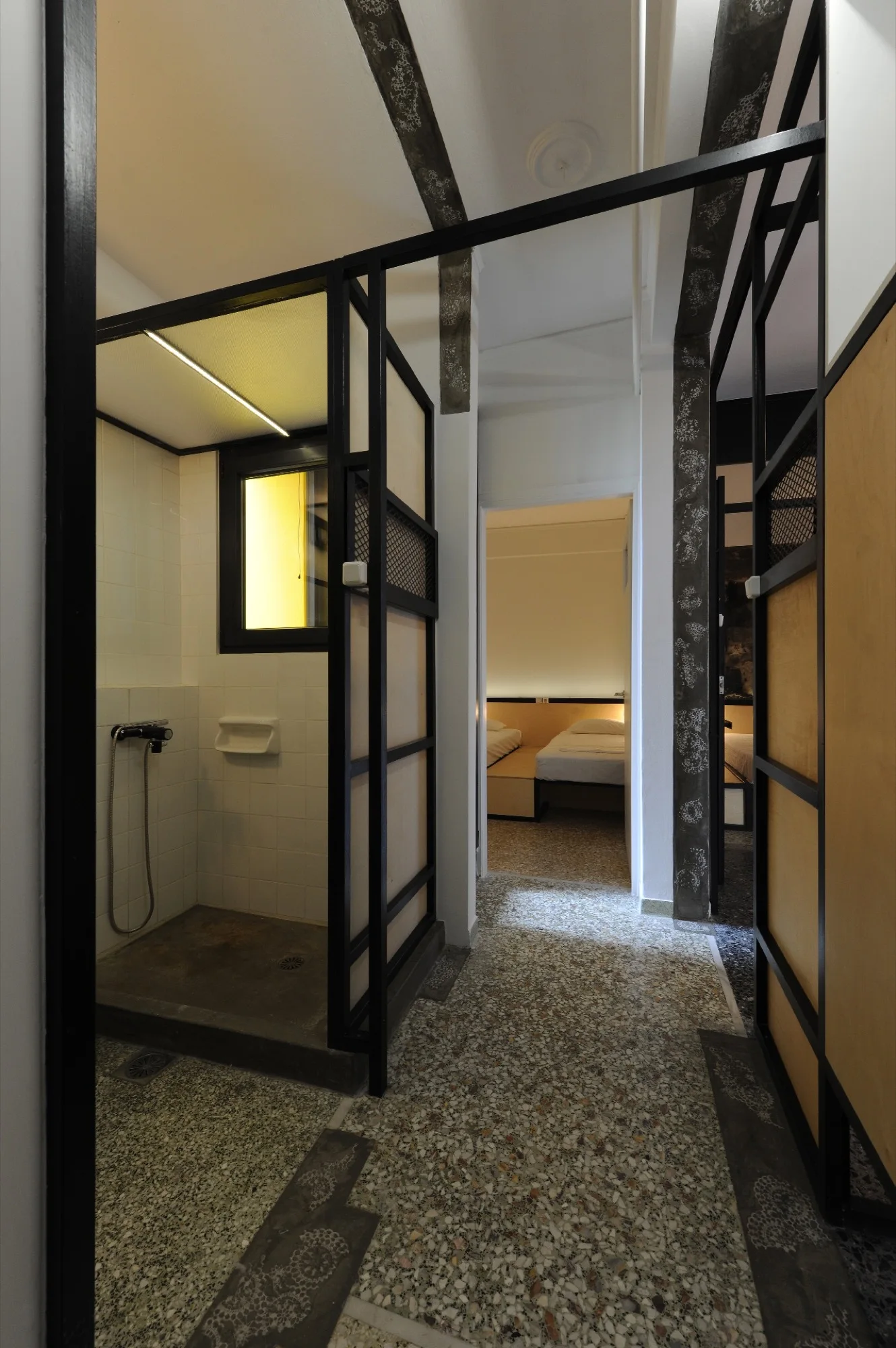 A hallway featuring speckled terrazzo flooring, a shower enclosed by black metal framed partitions, and an open doorway looking into a bedroom.