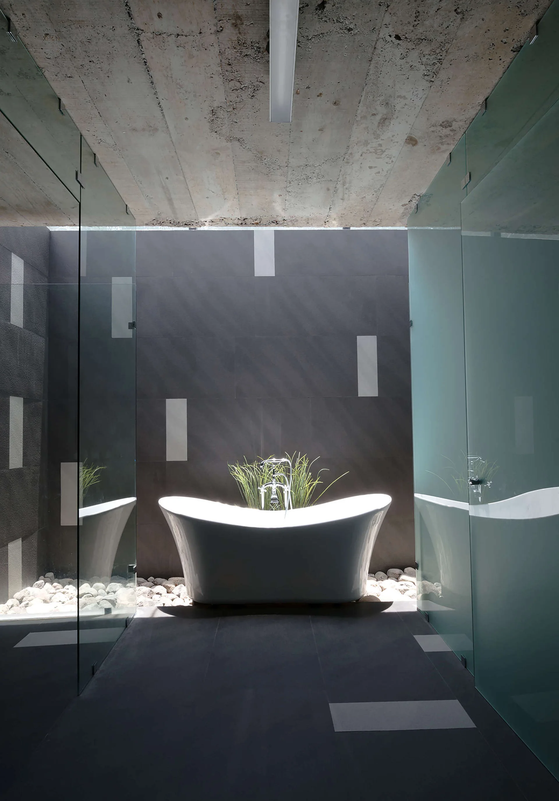 A white freestanding bathtub sits on a bed of white stones beneath an exposed concrete ceiling in a modern bathroom with dark tiled walls.