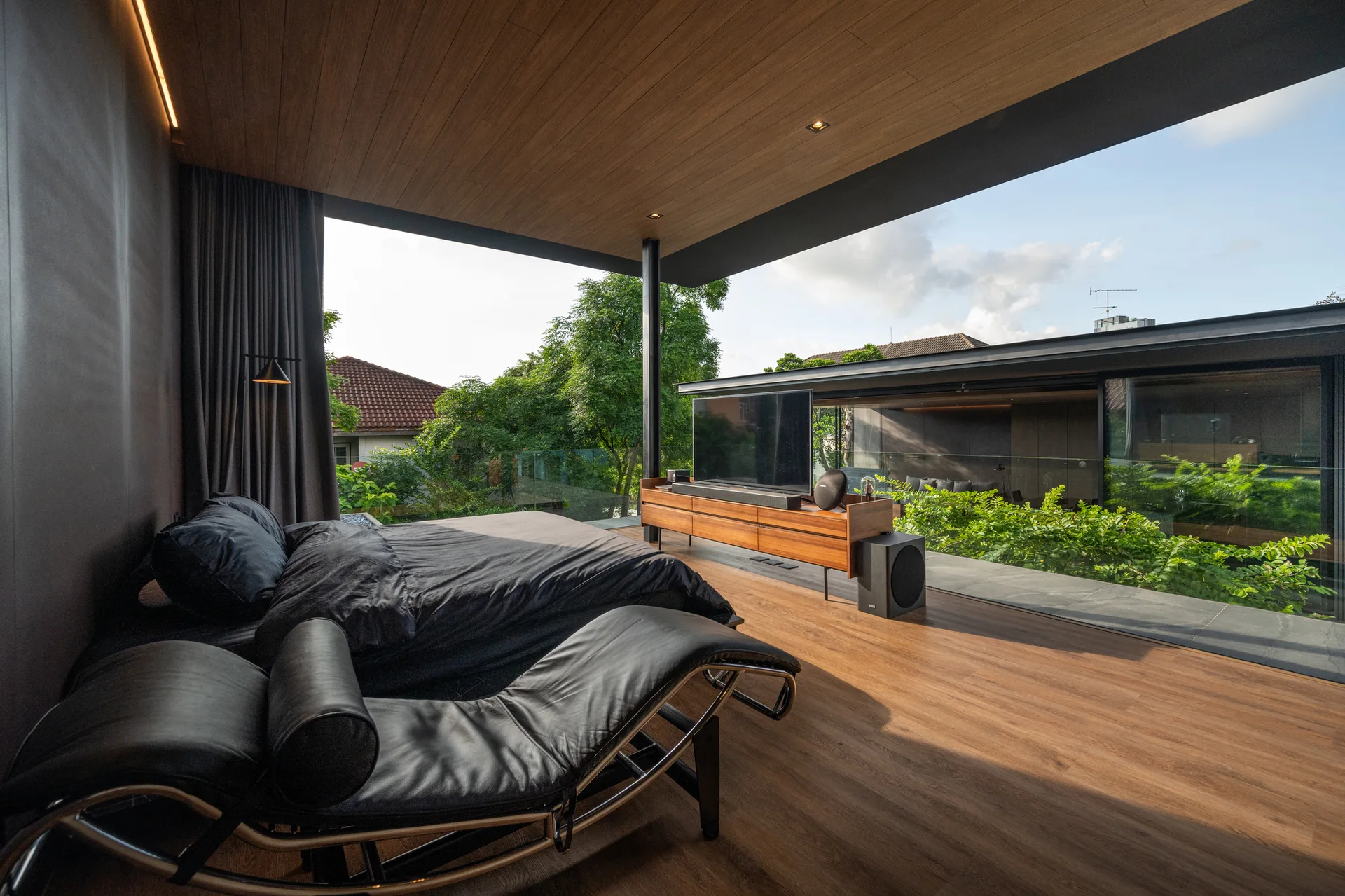 A minimalist bedroom with a black bed and chaise lounge features wood-paneled floors and ceilings that open directly onto an outdoor terrace.