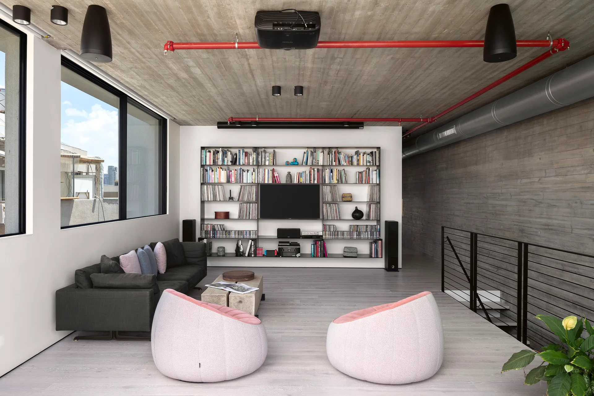 A modern industrial living room with an exposed concrete ceiling, a red sprinkler pipe, a black shelving unit, and two pink floor chairs.