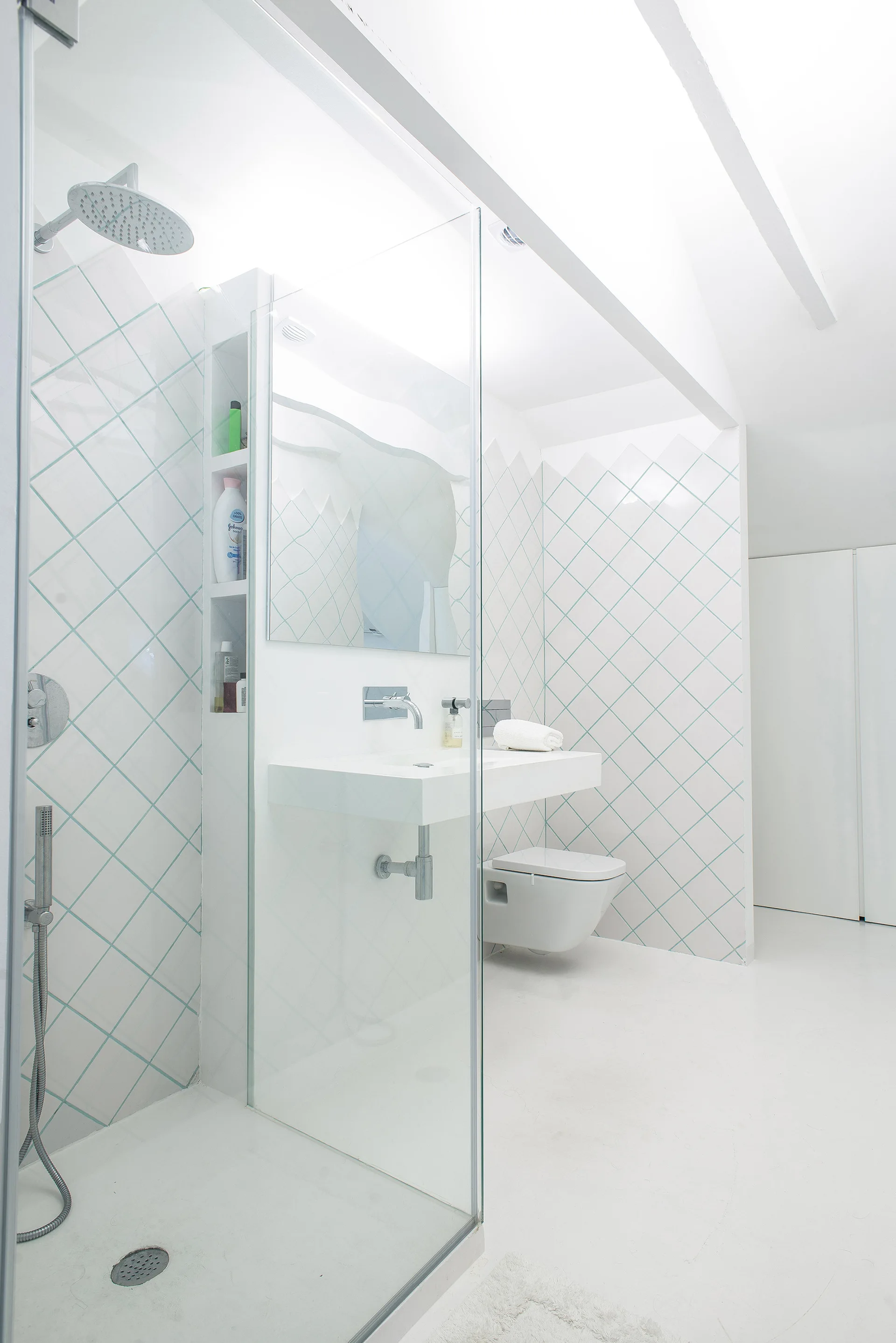 A minimalist white bathroom featuring a glass walk-in shower, a floating vanity, and white diamond-shaped wall tiles with turquoise grout.