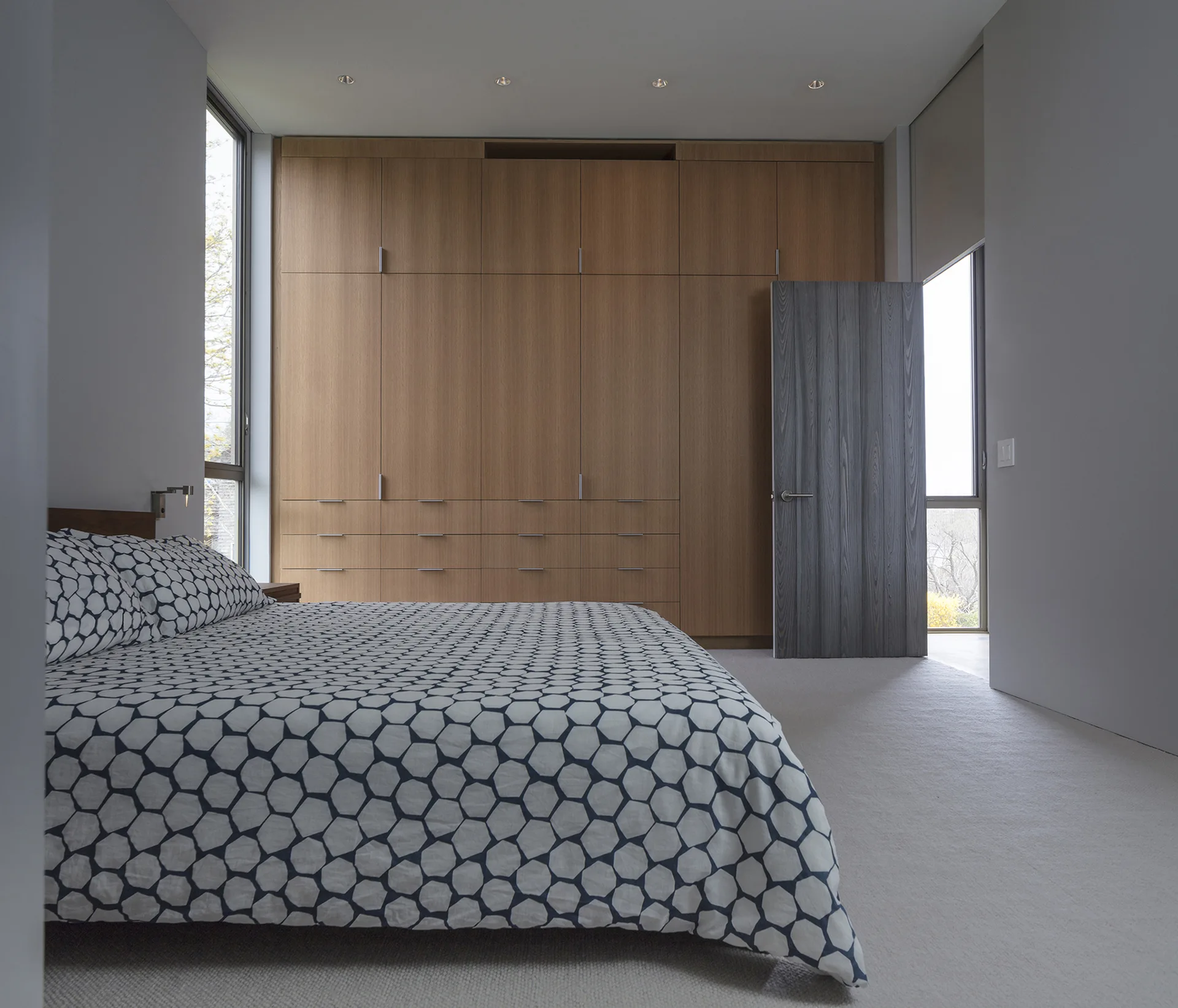 A minimalist bedroom featuring a bed with a geometric-patterned duvet, light grey walls, a grey wooden door, and a built-in wooden wardrobe wall.