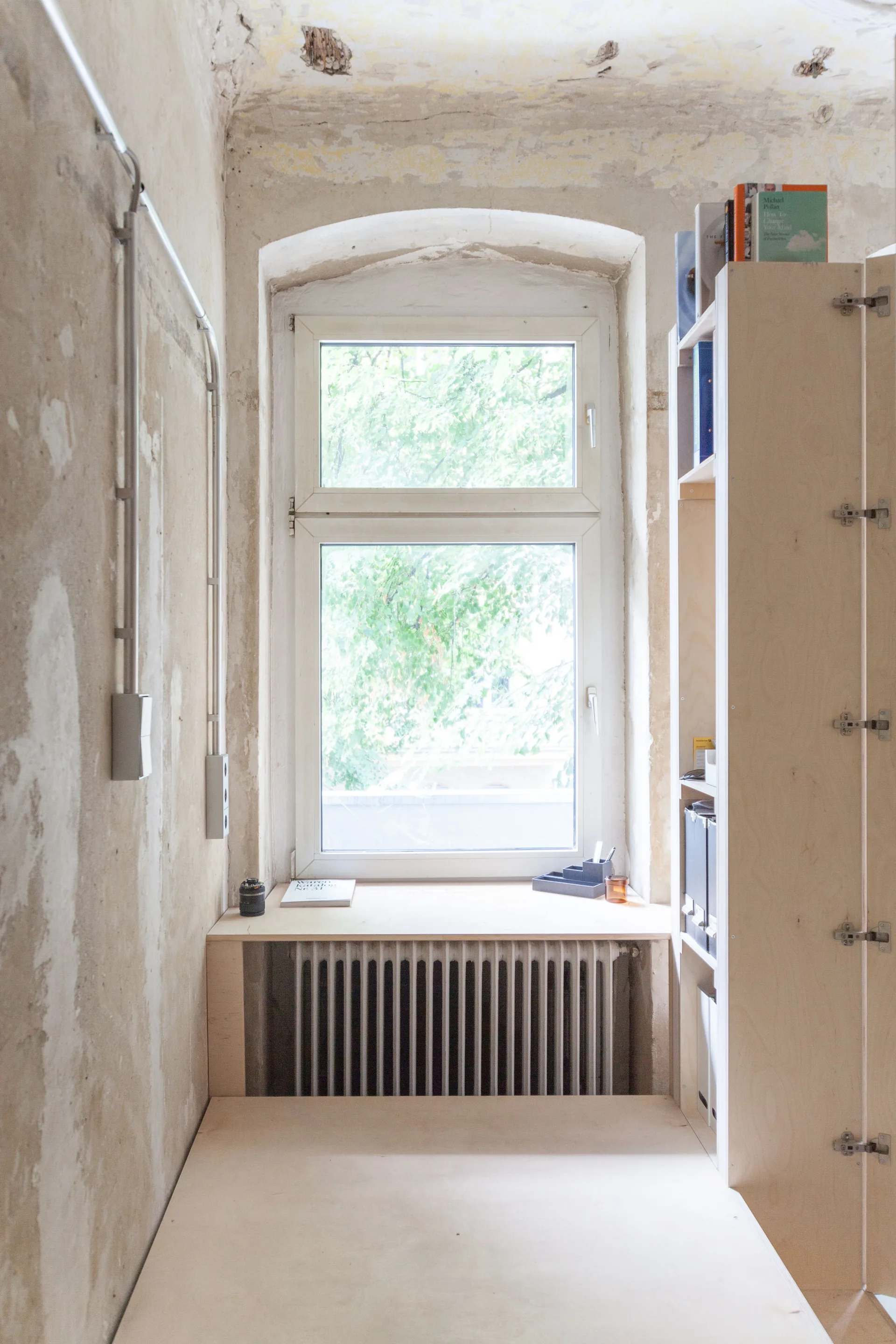 A custom plywood desk and built-in bookshelf stand next to an arched window and an exposed radiator against raw plaster walls.
