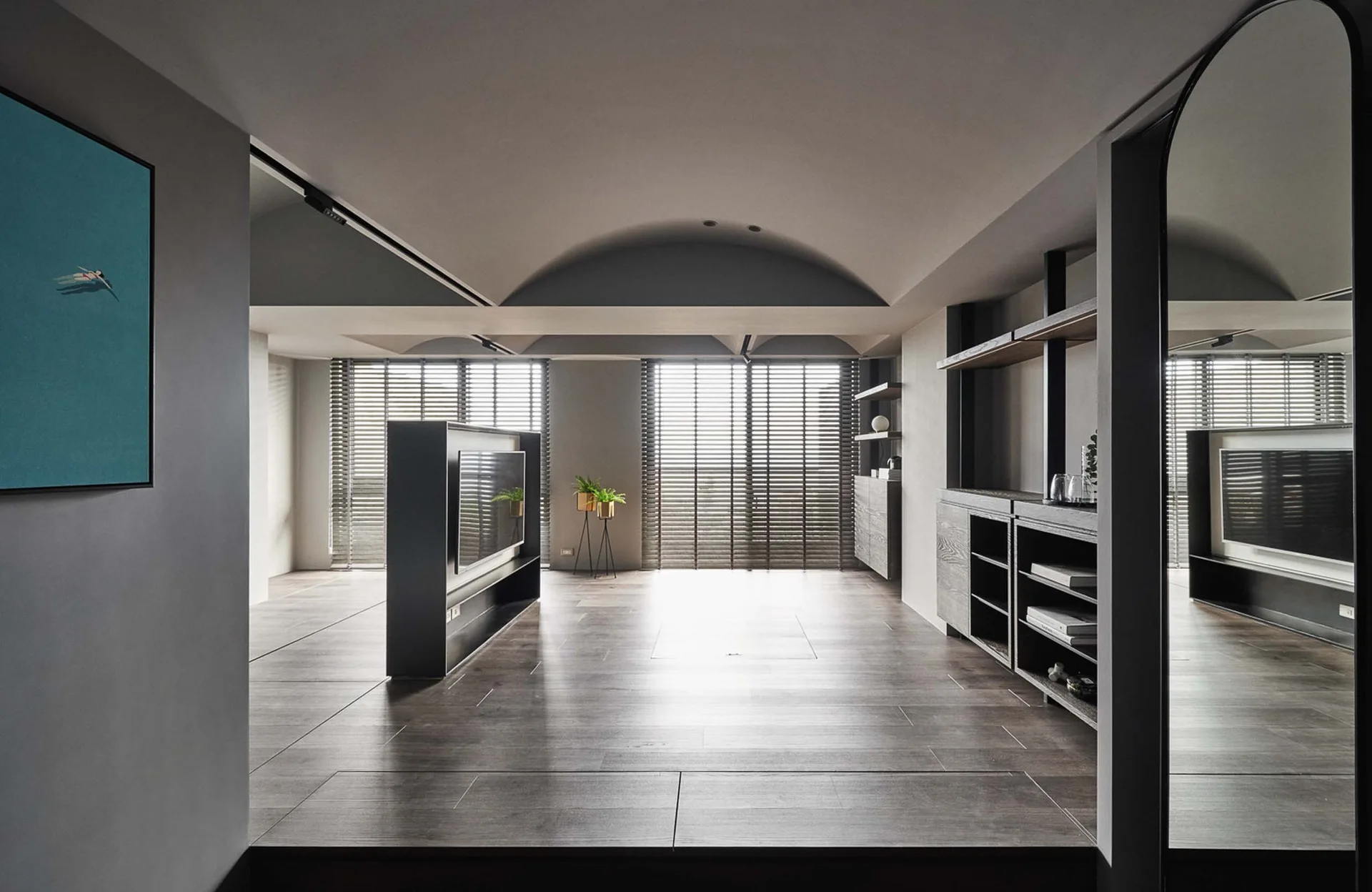 A minimalist grey living room featuring vaulted ceilings, dark wood flooring, a central freestanding TV partition, and windows with venetian blinds.