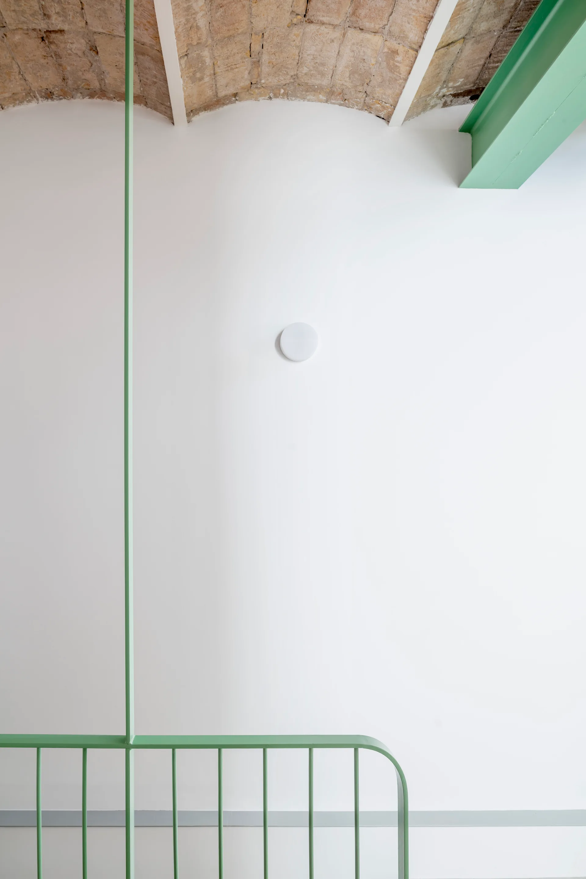 A minimalist interior featuring a sage green metal railing and painted beam against a white wall beneath an exposed brick vaulted ceiling.