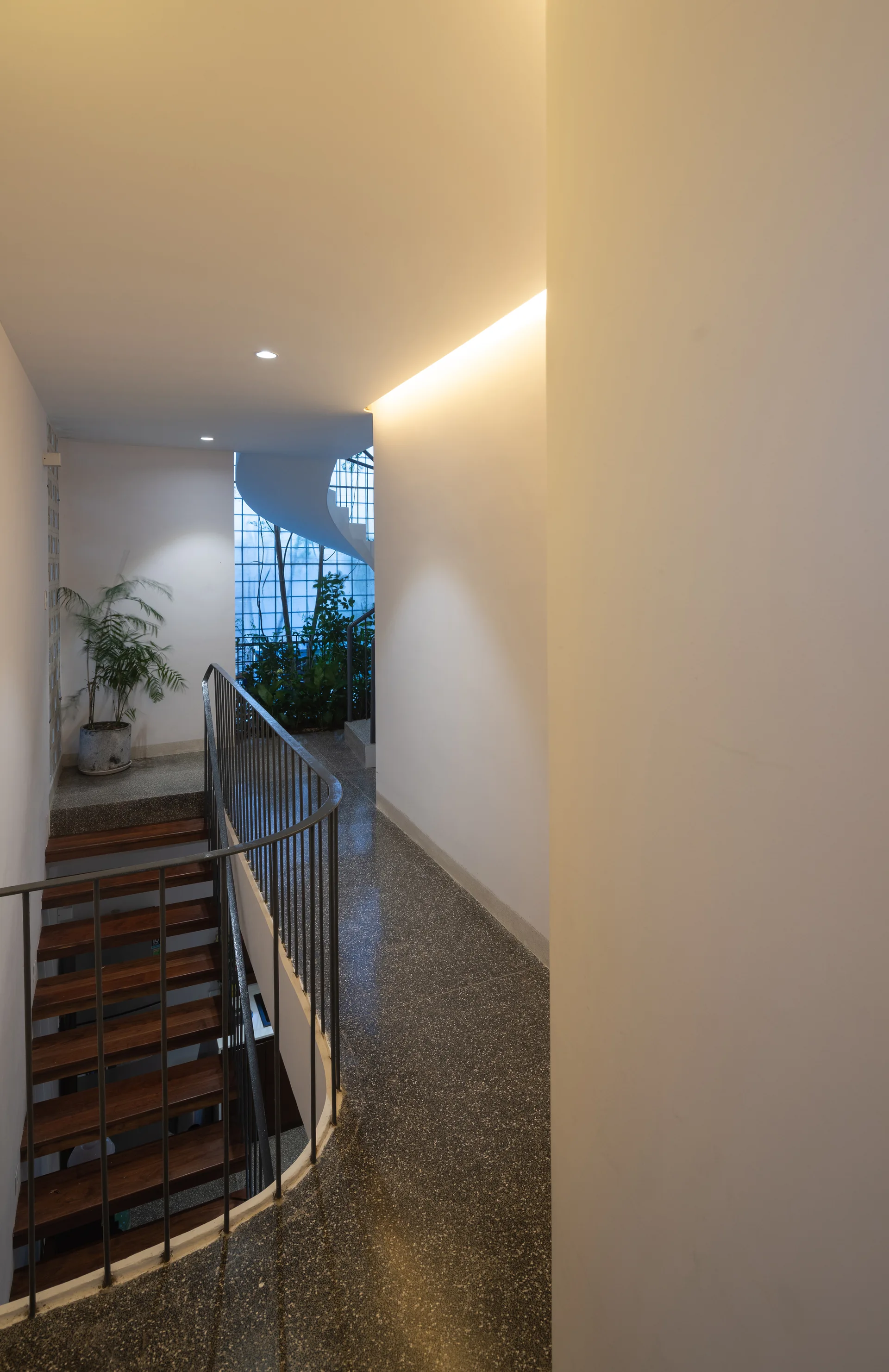 A curved staircase with wooden treads and a metal railing features a speckled grey floor and a white wall illuminated by warm ceiling cove lighting.