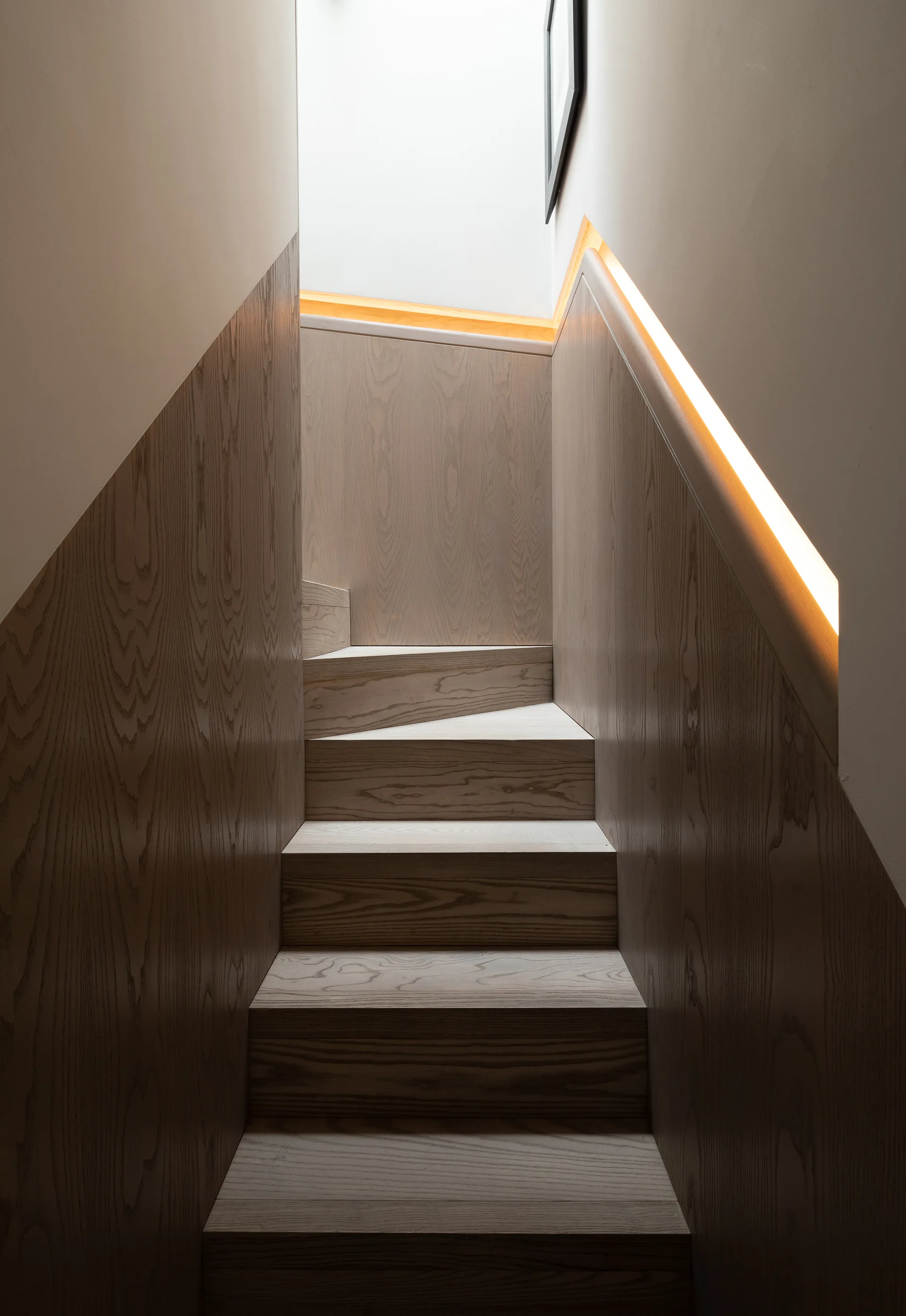 A modern wooden staircase with light-colored timber steps, matching wall paneling, and an integrated glowing warm LED strip light built into the wall.
