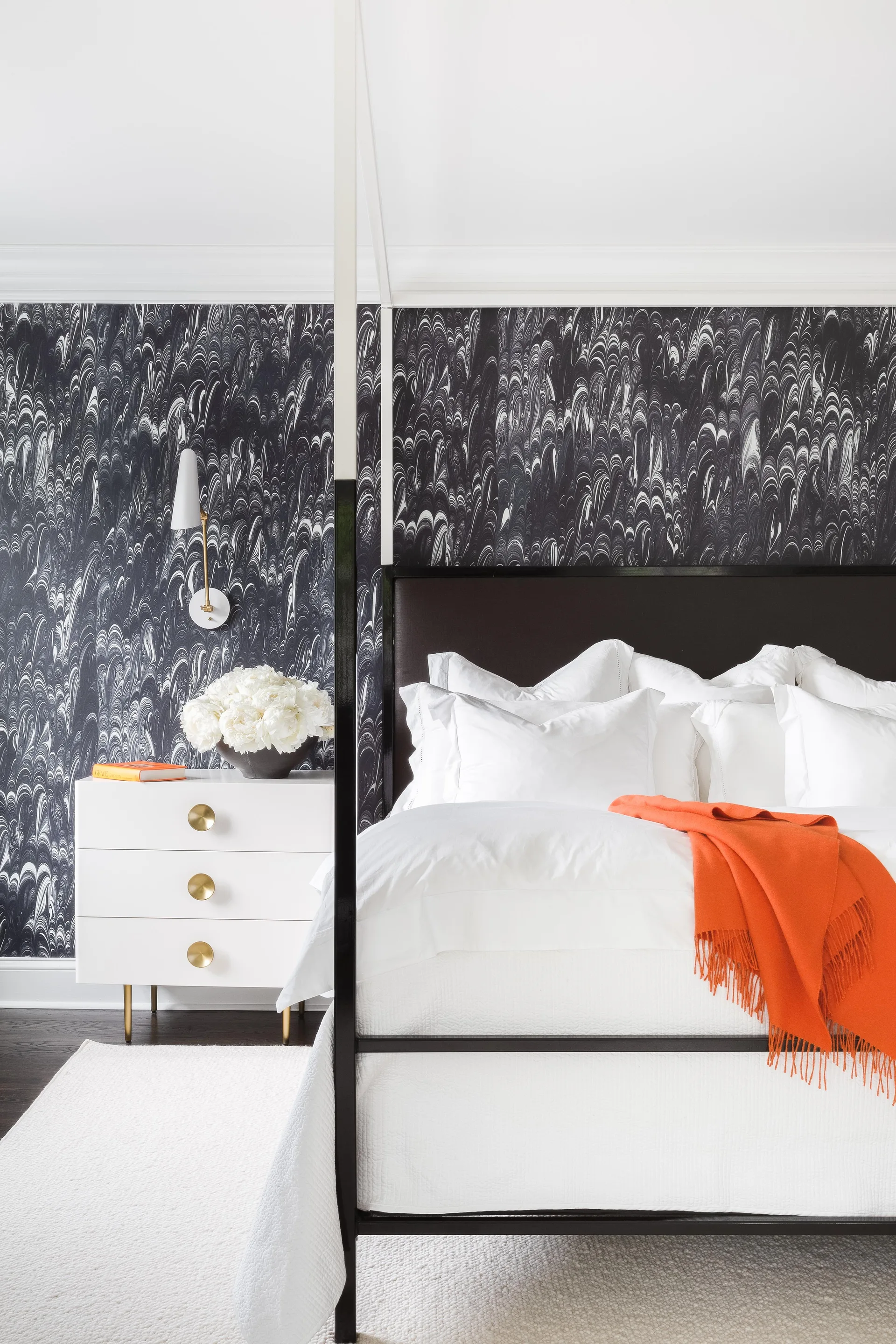 A modern bedroom features a black four-poster bed set against black and white marbled wallpaper next to a white three-drawer nightstand.