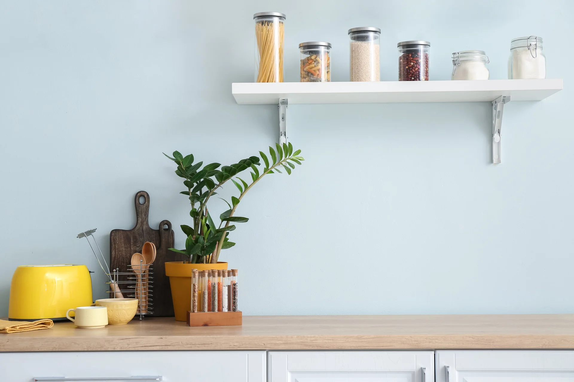 A white floating shelf with glass food jars is mounted on a light blue wall above a wooden kitchen countertop with a yellow toaster and green plant.