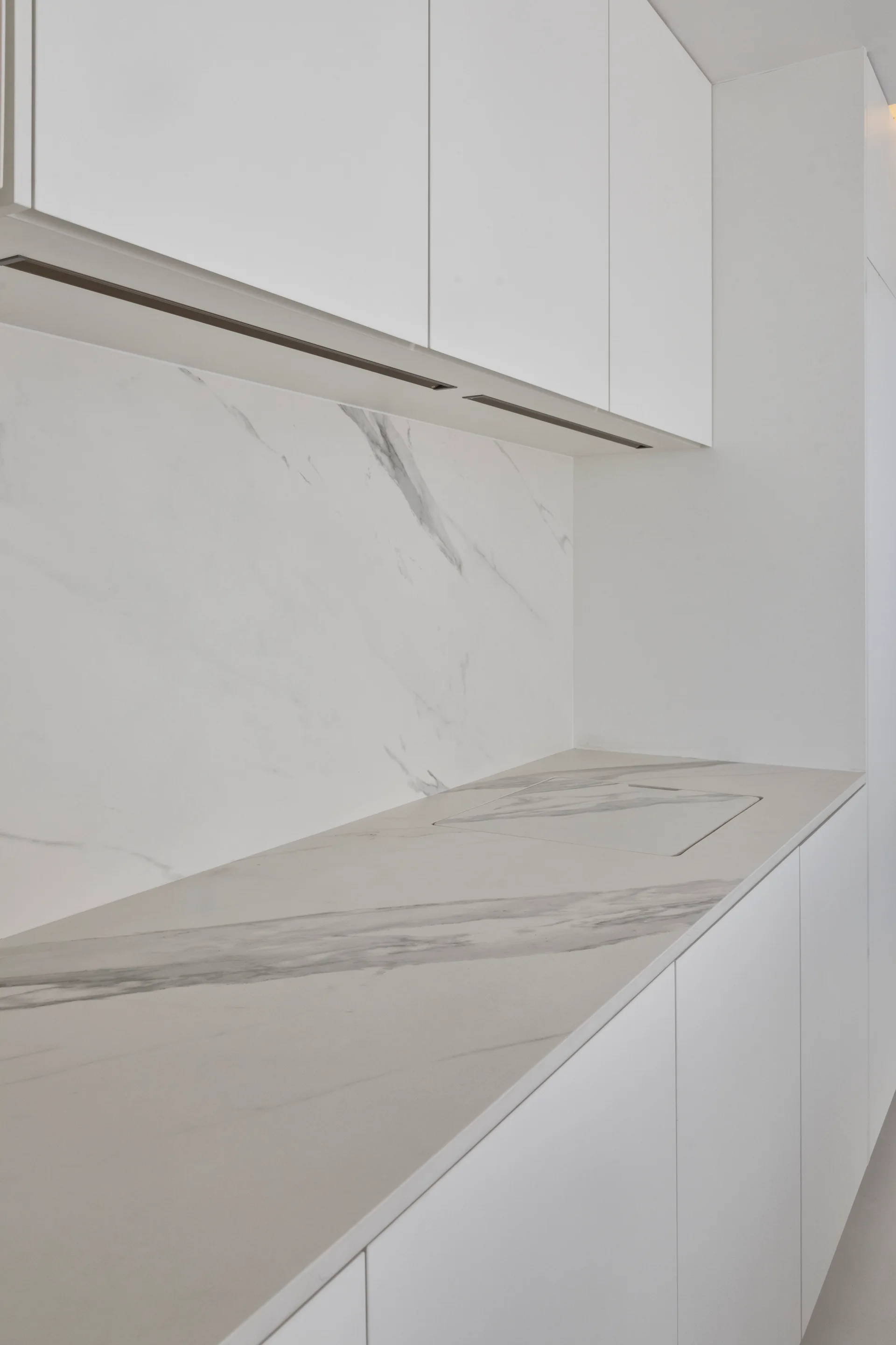 A minimalist white kitchen countertop with a veined stone slab backsplash and an integrated sink below flat-panel upper cabinetry.